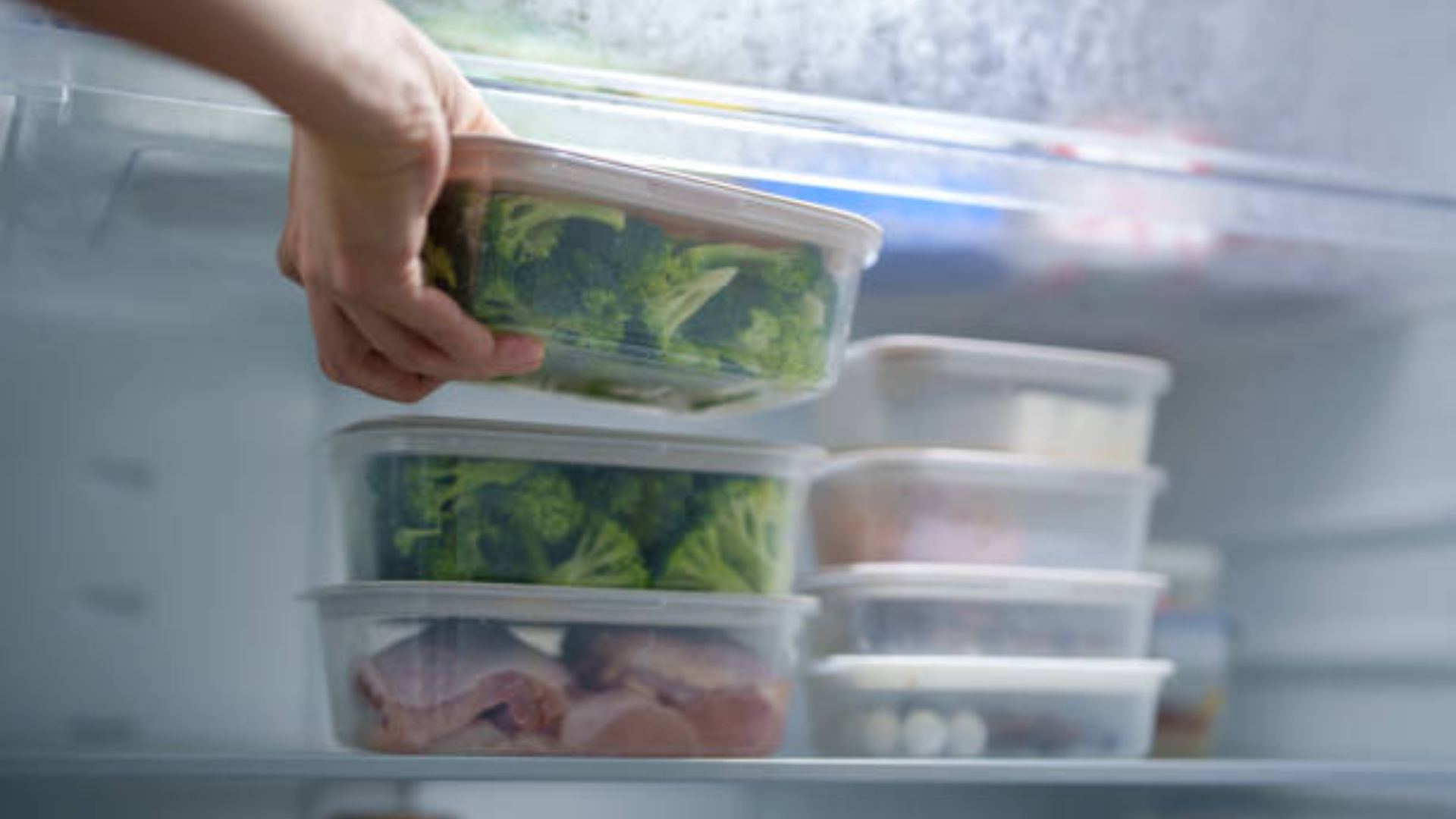 A close-up view of a hand placing a clear plastic container filled with fresh broccoli into a well-organized refrigerator alongside other food storage bins.