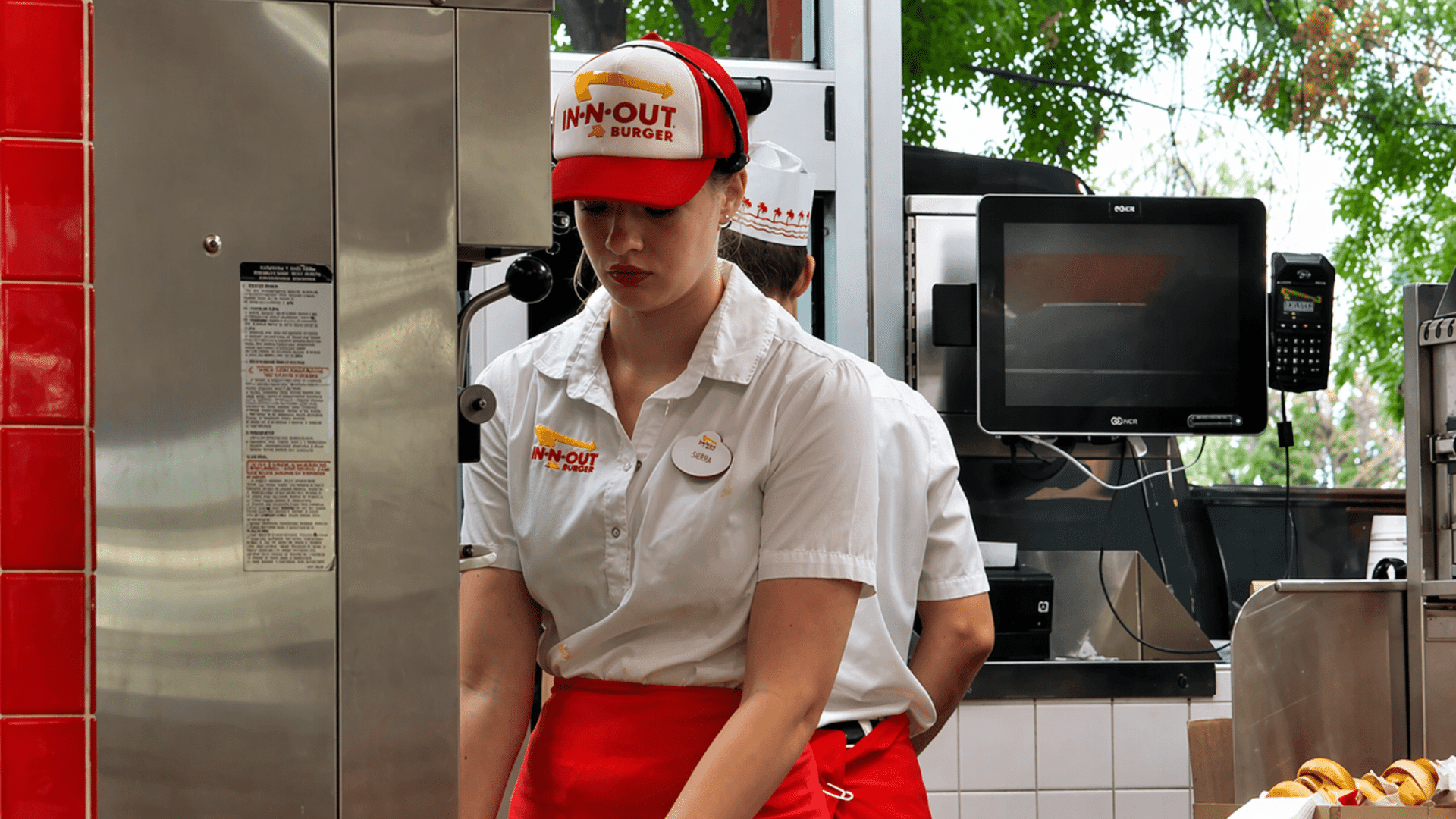 A medium shot of a female In-N-Out associate in a red-and-white ball cap and white uniform, looking down while working at a stainless steel station.
