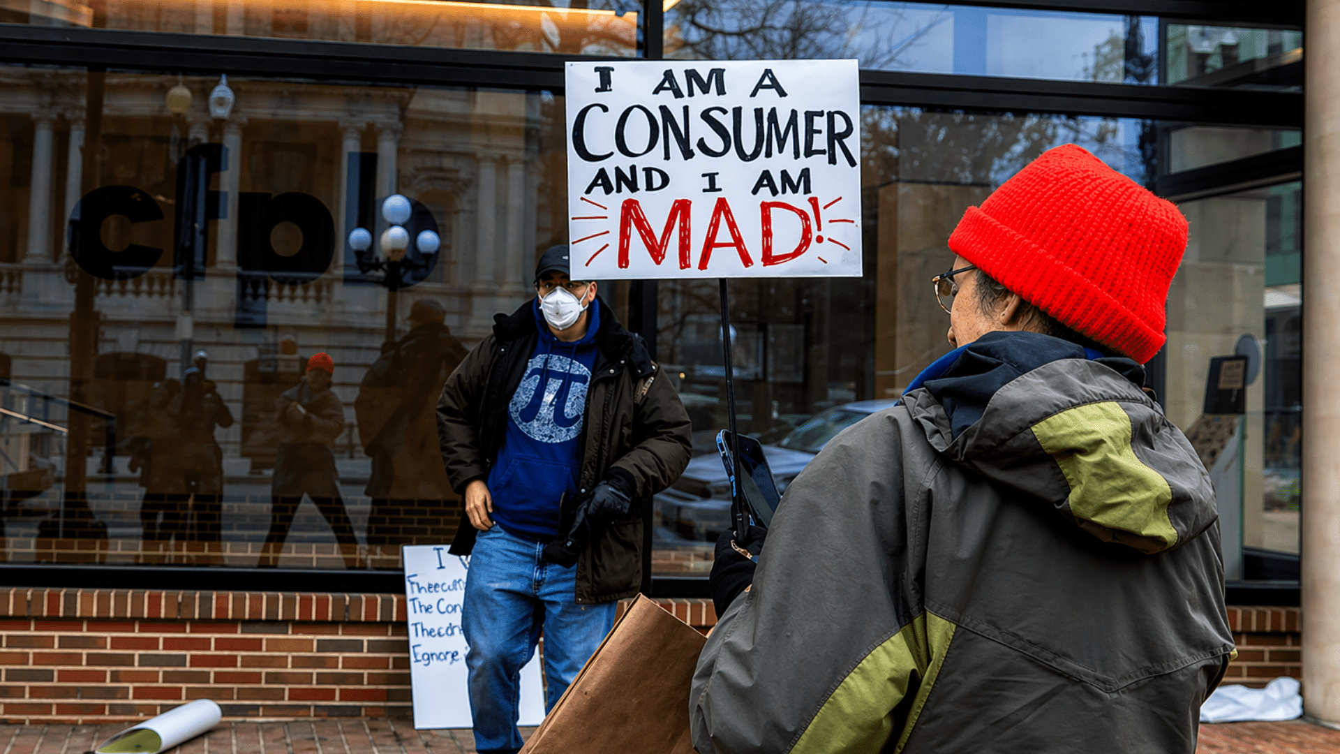 A person in a red beanie holds up a sign that reads "I AM A CONSUMER AND I AM MAD!" while another person in a face mask stands nearby.