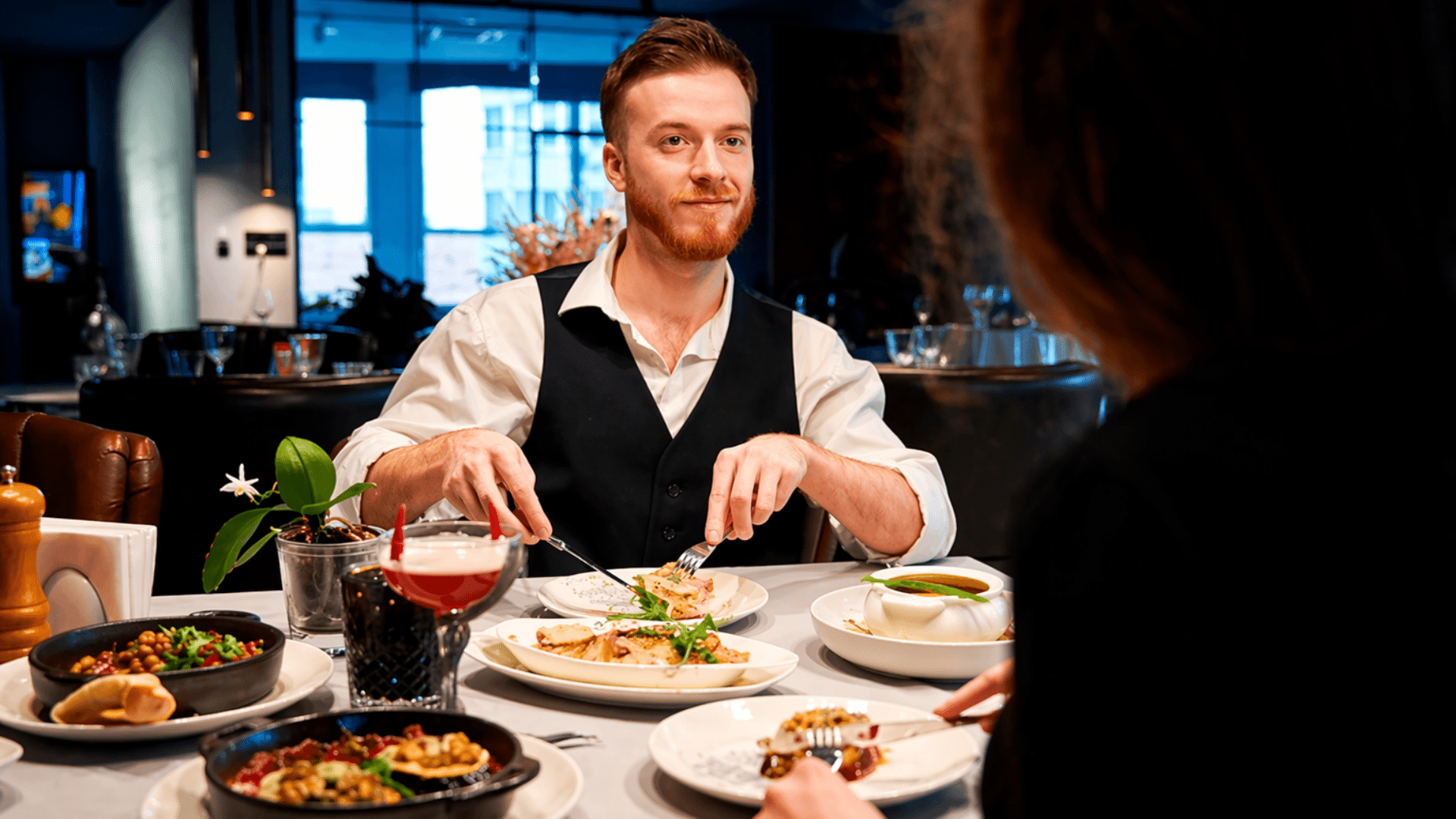 A medium shot of a young man with a red beard, wearing a white shirt and black vest, dining at a modern restaurant; he is looking across the table at his companion while cutting his food.