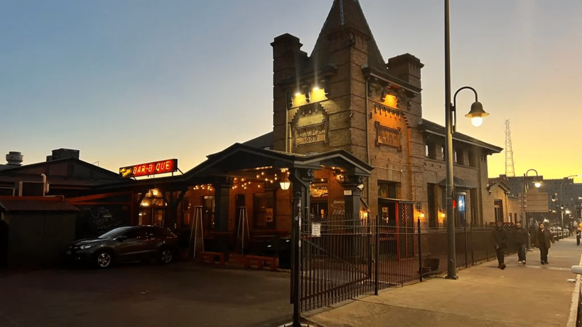 A scenic shot of the Rochester Dinosaur Bar-B-Que at dusk; the restaurant is housed in the historic Lehigh Valley Railroad Station, with warm yellow lighting illuminating the stone tower and a "BAR-B-QUE" neon sign visible on the left.