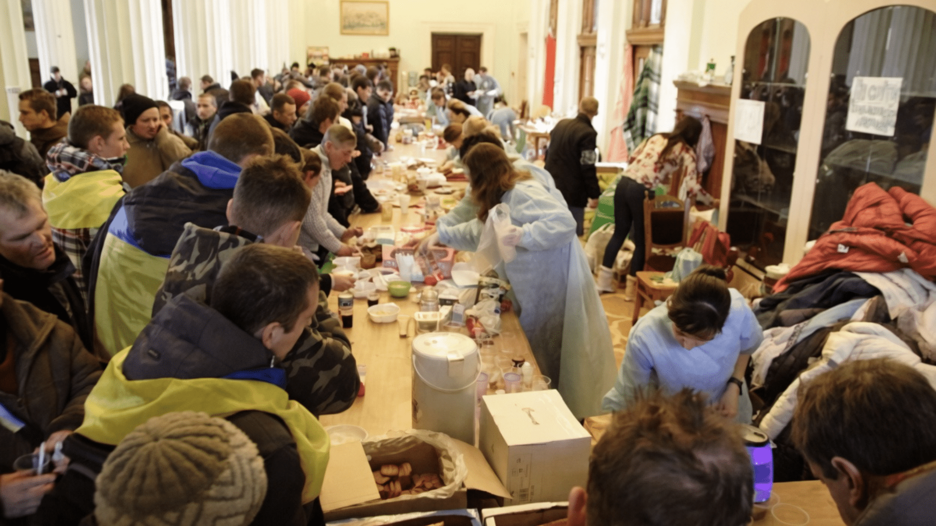 A wide indoor view of a crowded humanitarian aid center where volunteers in blue and yellow protective gear serve meals to a large group of people at long tables.