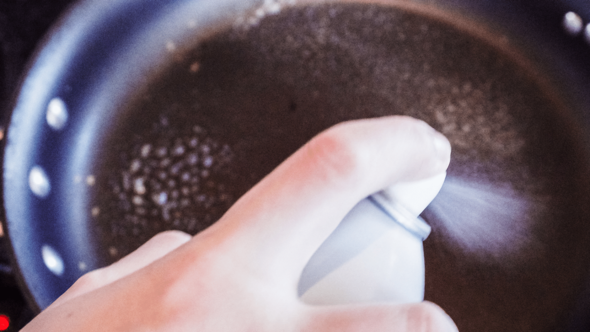 A hand sprays cooking oil from a white can into a dark frying pan.