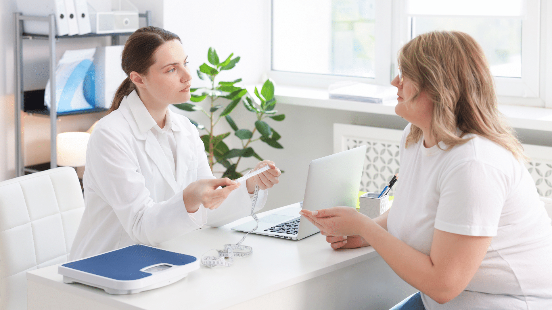A doctor holding a measuring tape sits at a desk talking with a female patient.