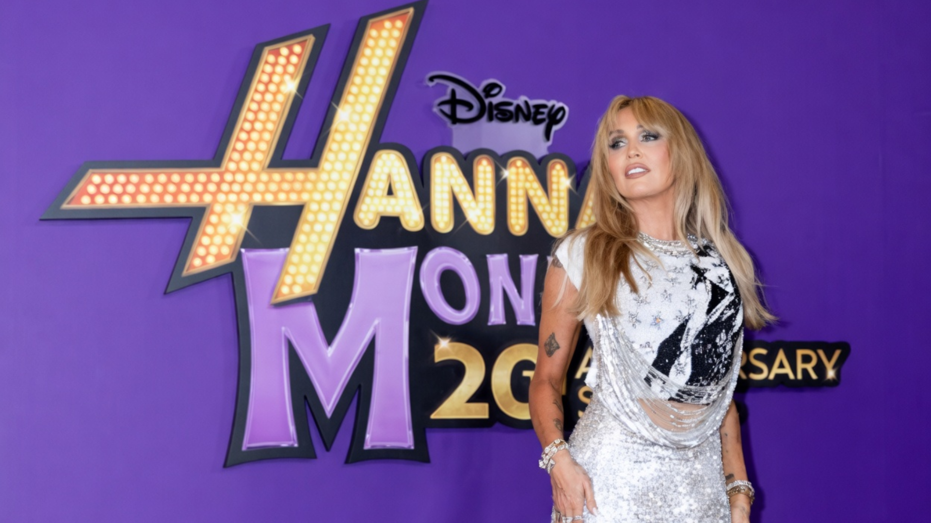 Miley Cyrus in a sparkling silver dress posing in front of a purple backdrop featuring the "Hannah Montana 20th Anniversary" logo.