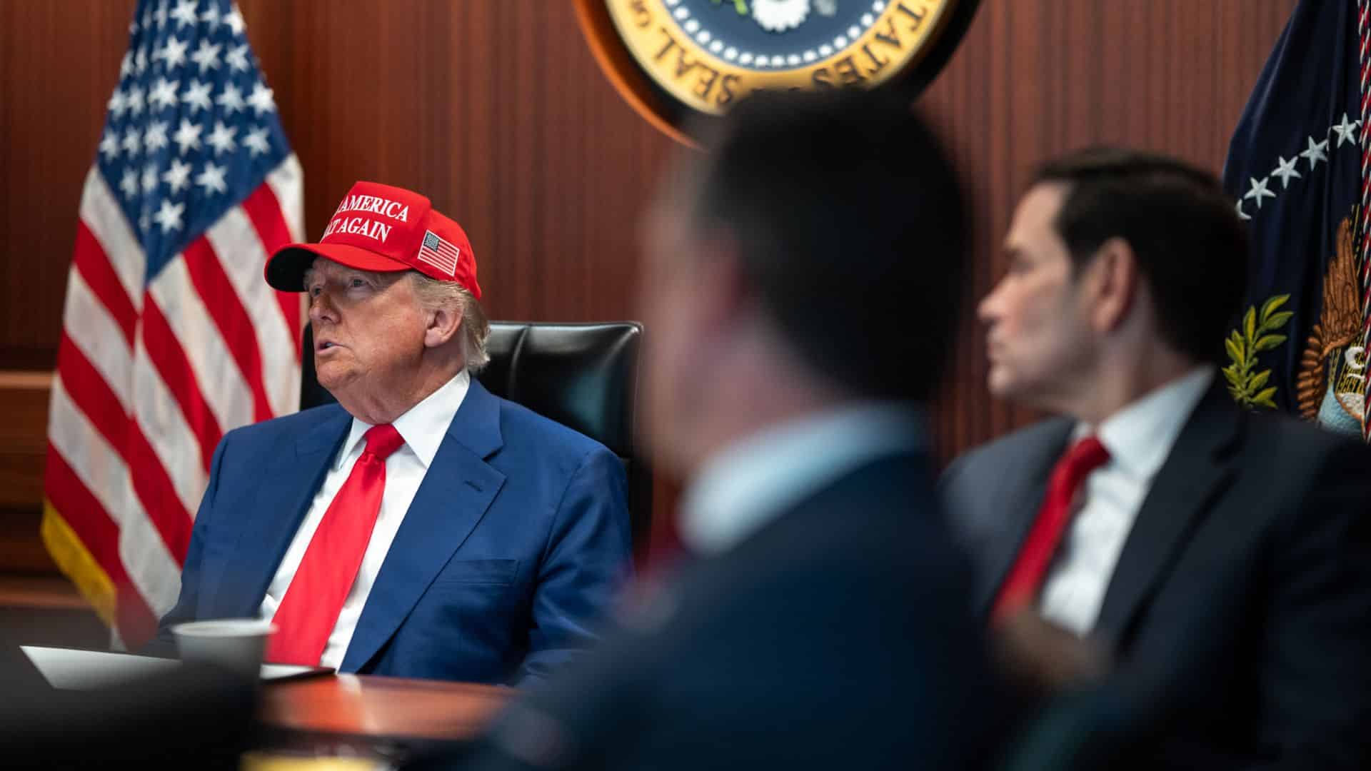 President Trump, in his red MAGA hat and blue suit, speaks during a meeting room discussion.
