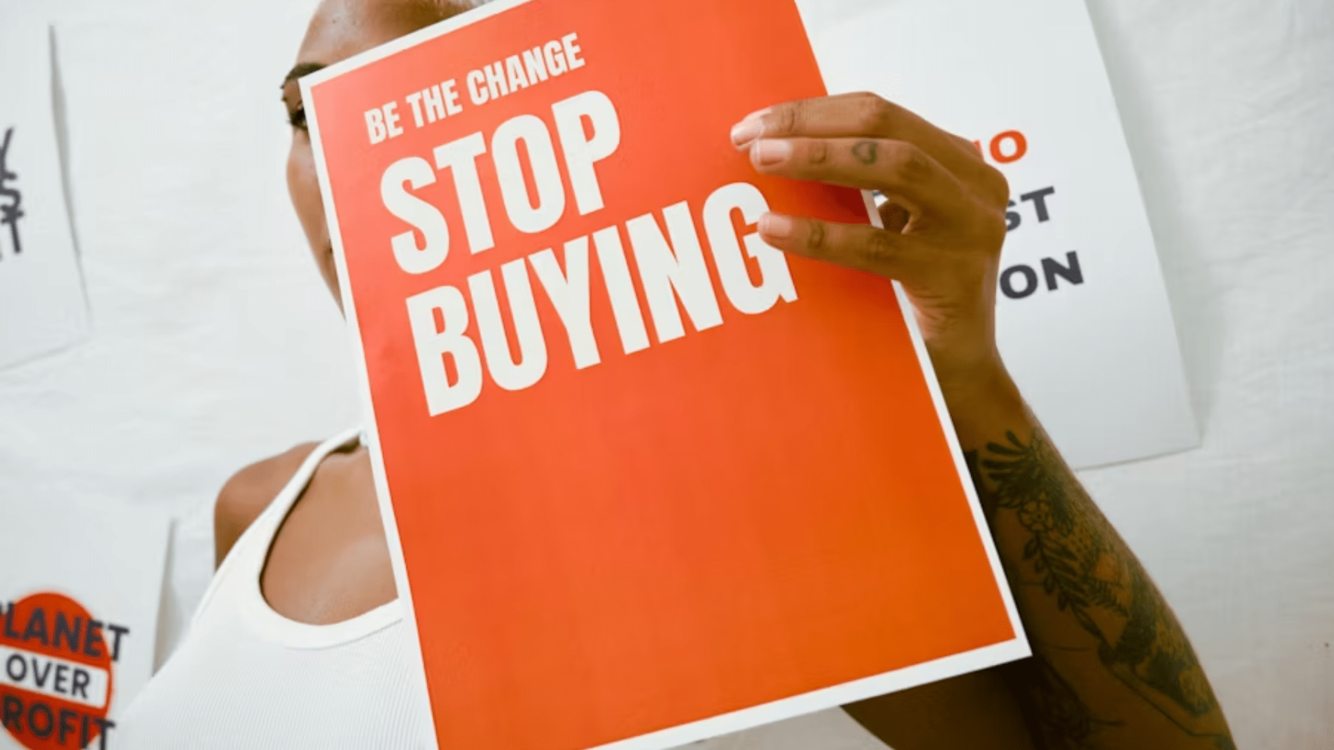 A close-up of a person holding an orange-and-white sign that reads "BE THE CHANGE STOP BUYING".