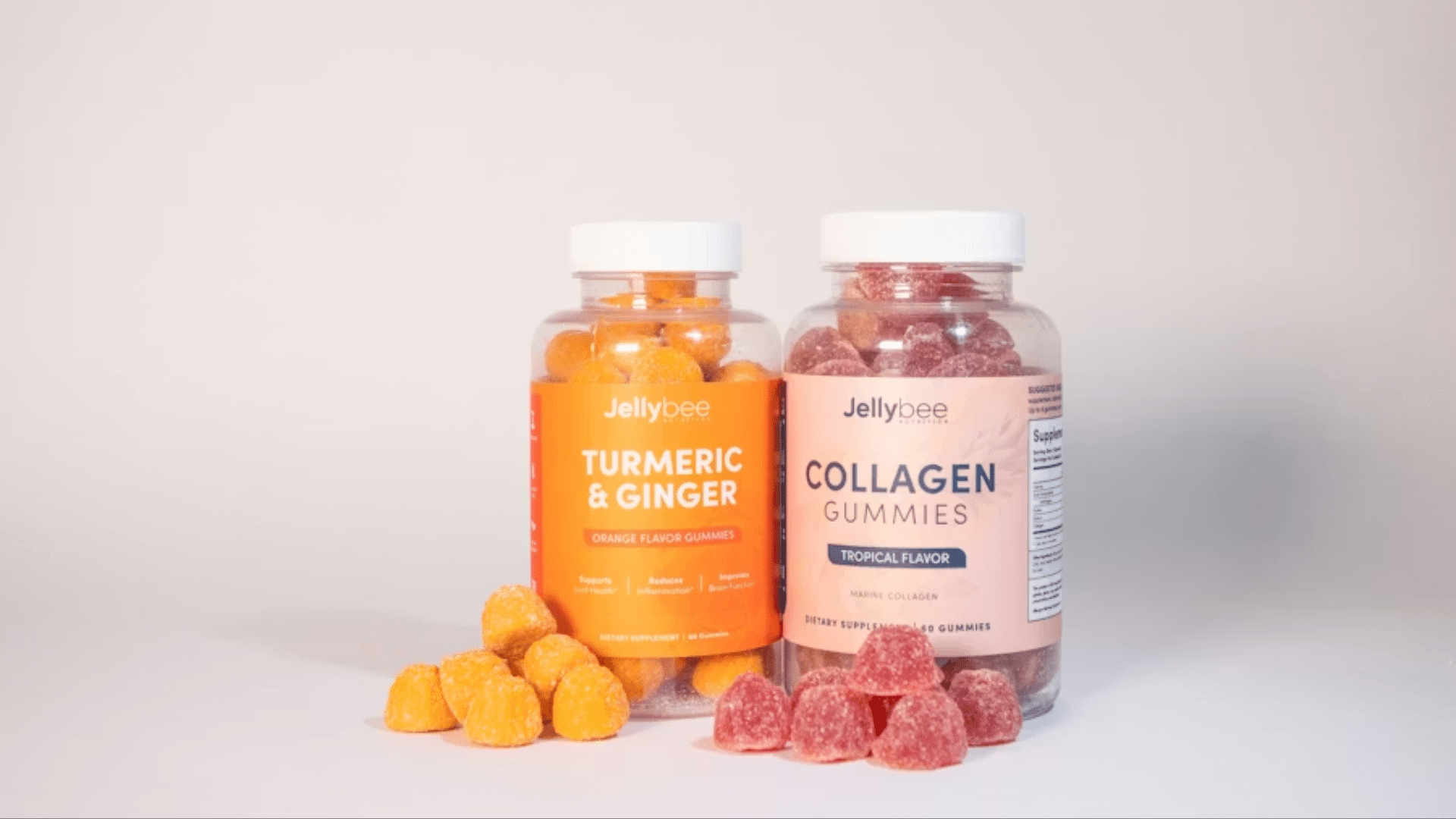 A studio product shot of two clear plastic jars of Jellybee Nutrition gummies on a white surface: one orange jar for "Turmeric & Ginger" and one pink jar for "Collagen Gummies," with loose gummies piled in front.