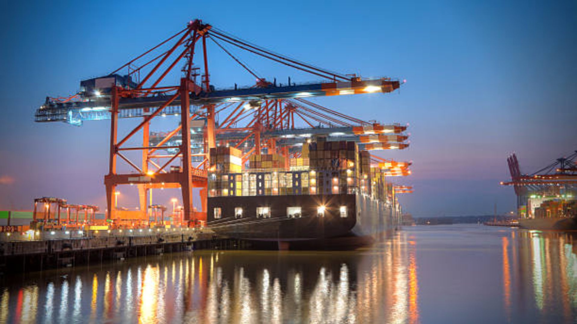 A nighttime view of a massive container ship docked at a port, with giant orange gantry cranes illuminated by bright lights while loading cargo.