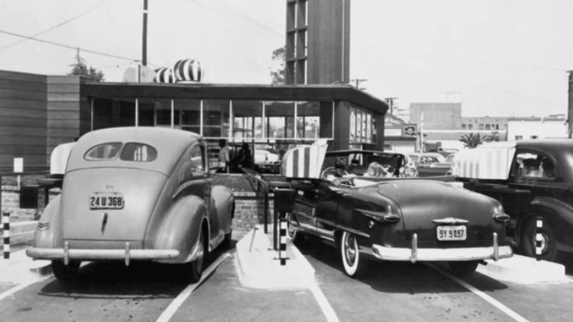 A vintage black-and-white photograph of the original In-N-Out Burger stand in Baldwin Park, California, showing two classic 1940s-era cars parked in the drive-thru lanes.