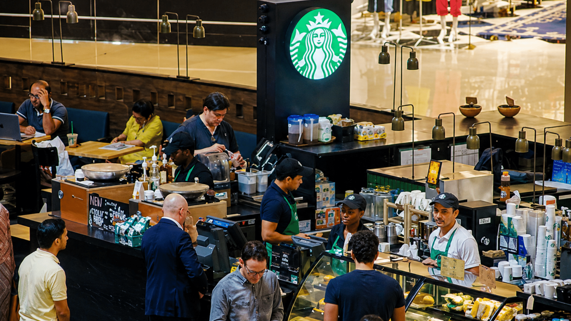 A high-angle shot looking down at a busy Starbucks location inside a shopping mall; multiple employees in green aprons are serving customers at the counter, while patrons sit at tables in the adjacent dining area.