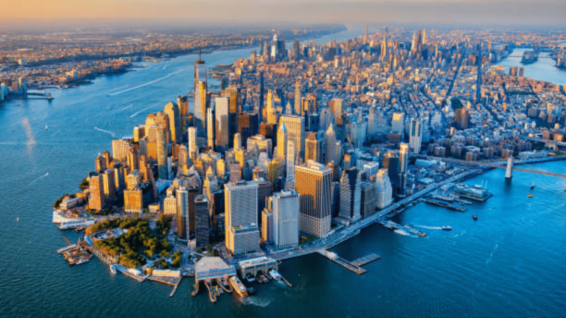 A sweeping aerial panorama of Manhattan Island at sunset, showing the dense skyscrapers of the Financial District and Midtown.