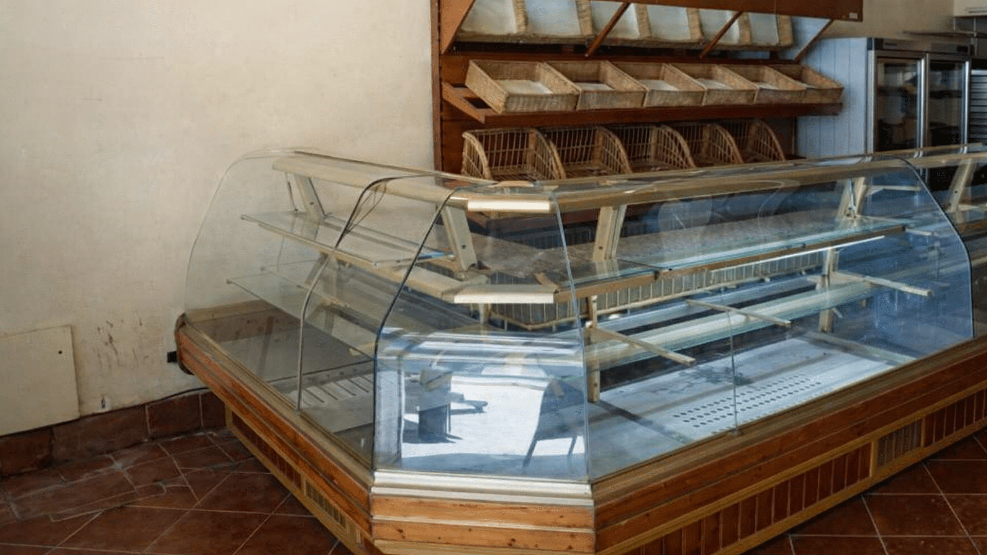 The interior of an empty bakery showing a curved glass display case with nothing inside and empty wooden bread racks against a beige wall.