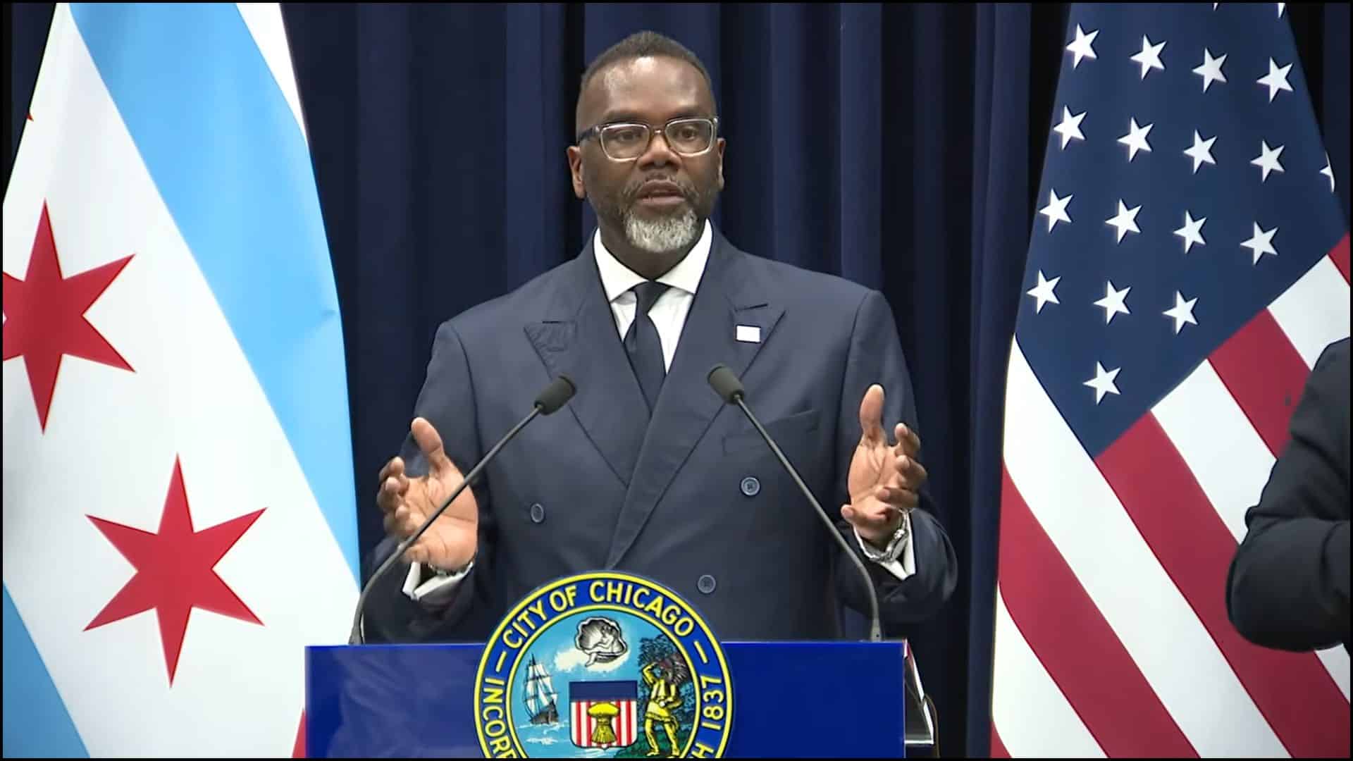Chicago Mayor Brandon Johnson gesturing while speaking at a press conference with U.S. and Chicago flags in the background.