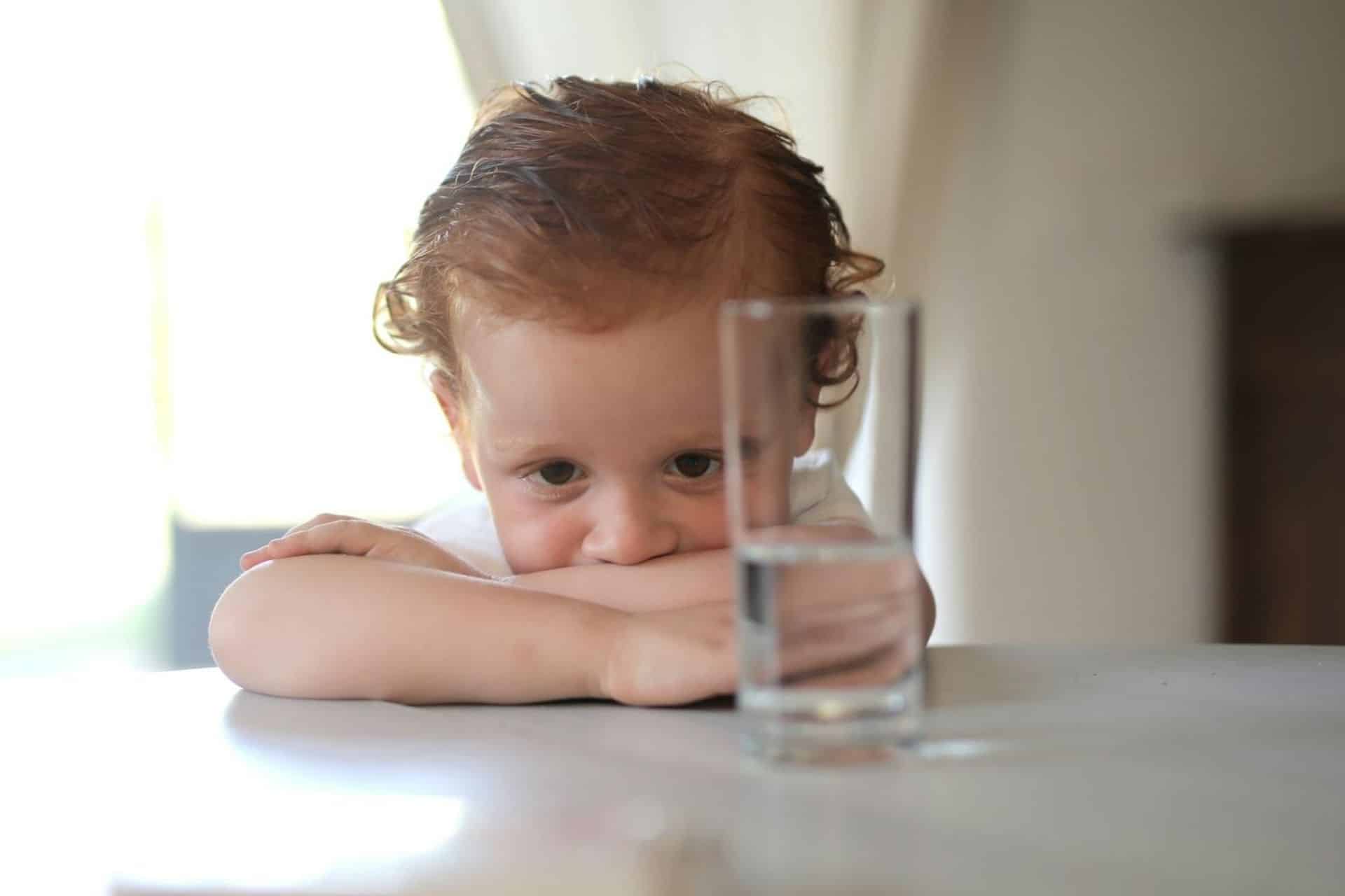 Young boy resting head on folded arms while looking at a glass of water on a table.