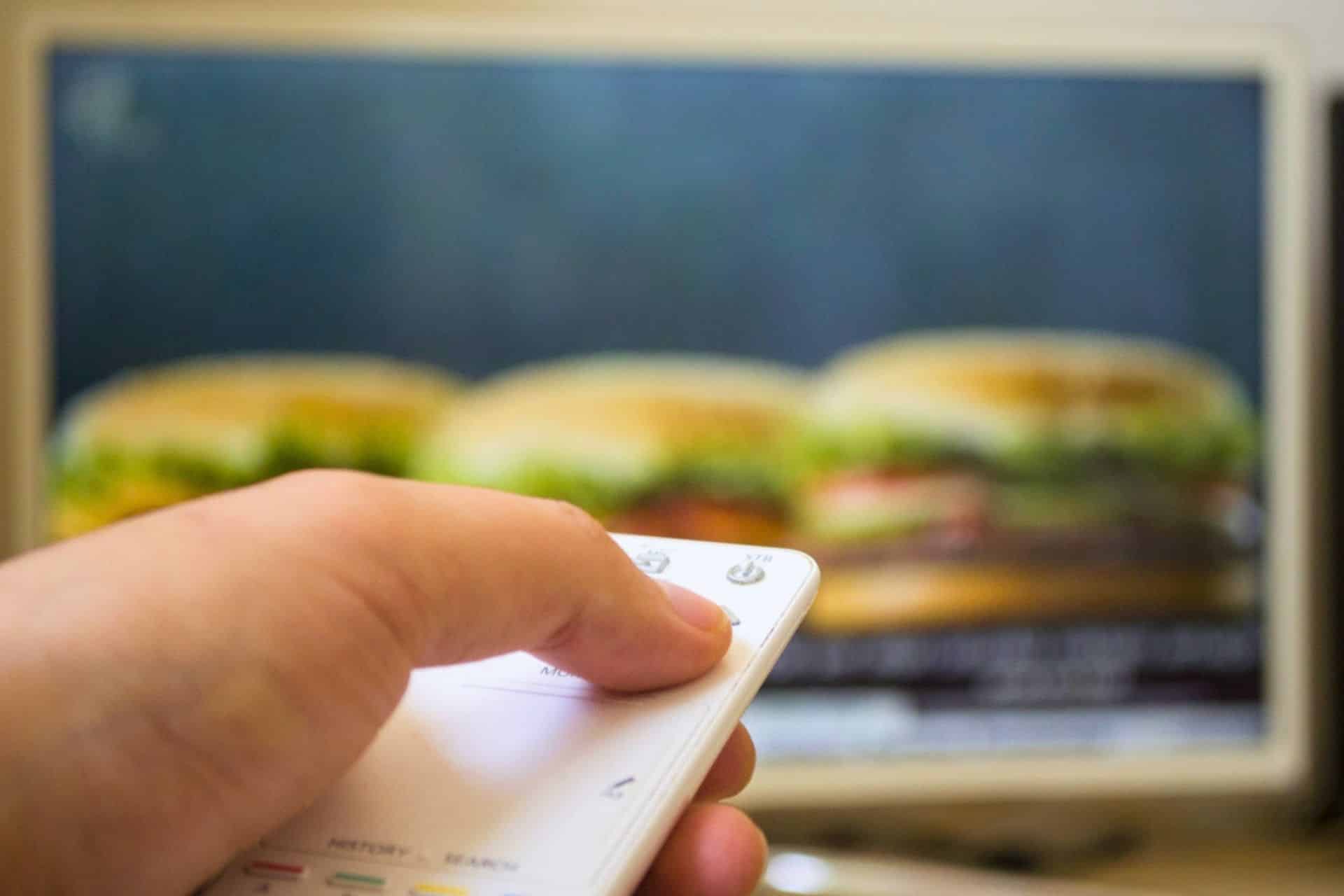Hand holding remote control pointed at TV screen showing blurred burger advertisement.