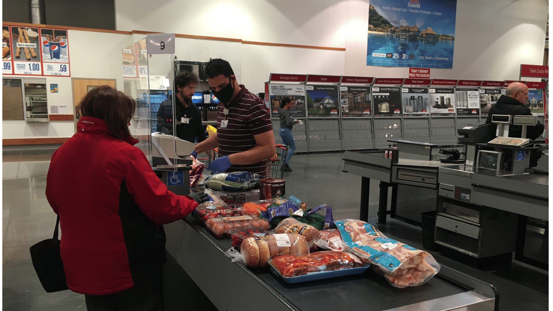 A cashier wearing a face mask and gloves scans a variety of bulk groceries at a warehouse club checkout counter.