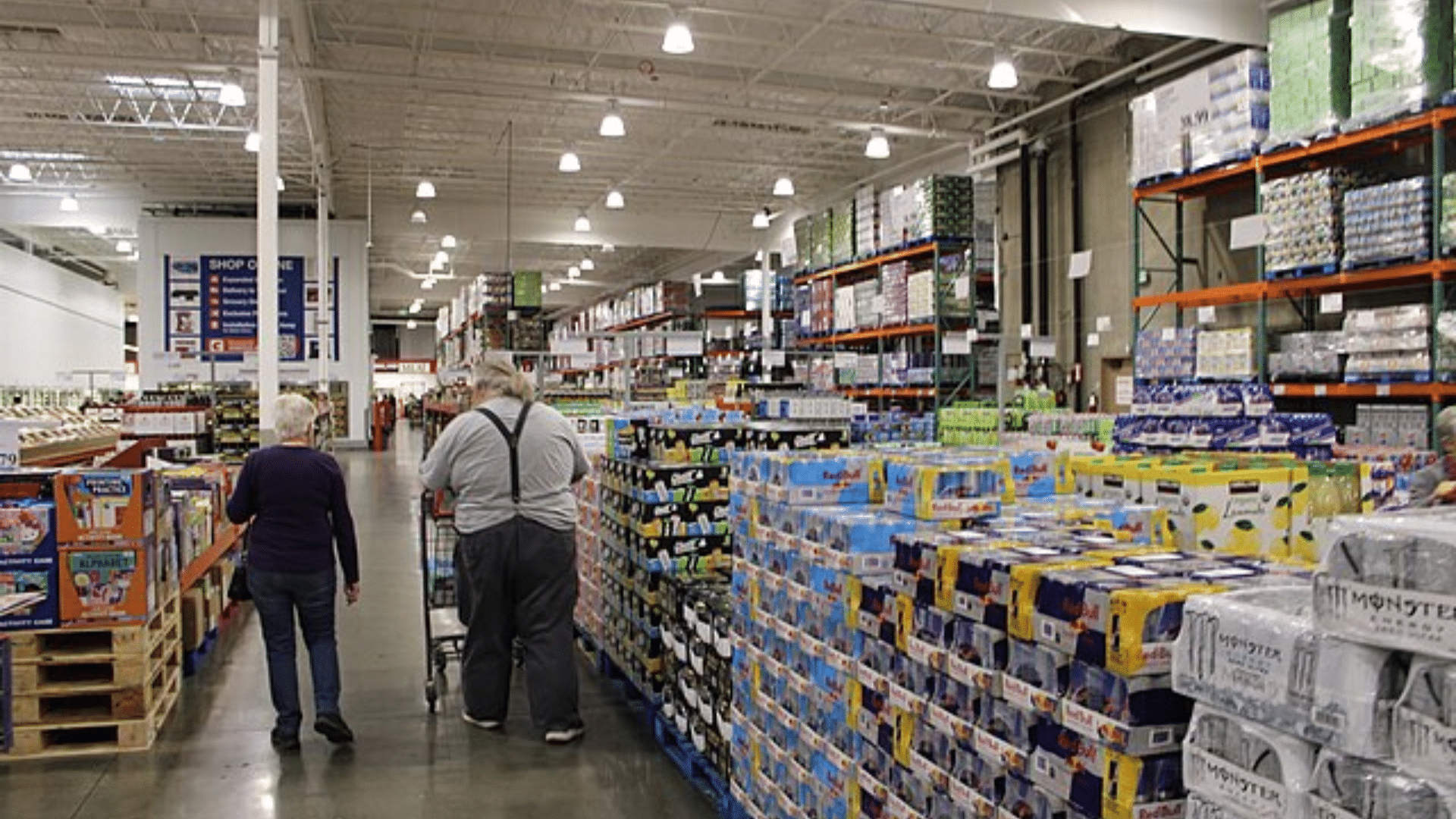 An interior view of a Costco warehouse showing customers walking down a wide aisle lined with bulk pallets of energy drinks and household goods.