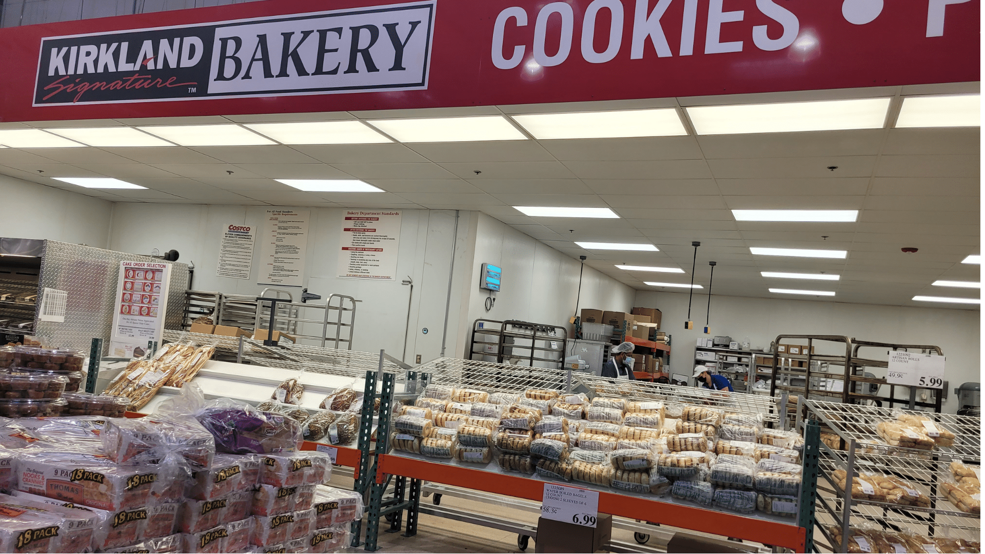 Shelves in a bakery department are stocked with various packaged breads, muffins, and cookies under a large red sign.