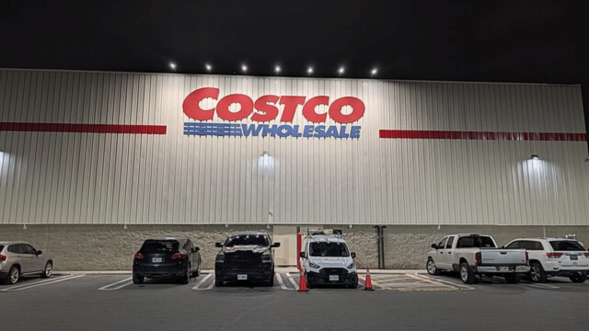 The exterior of a Costco Wholesale warehouse building at night with its large red logo illuminated above a row of parked cars.
