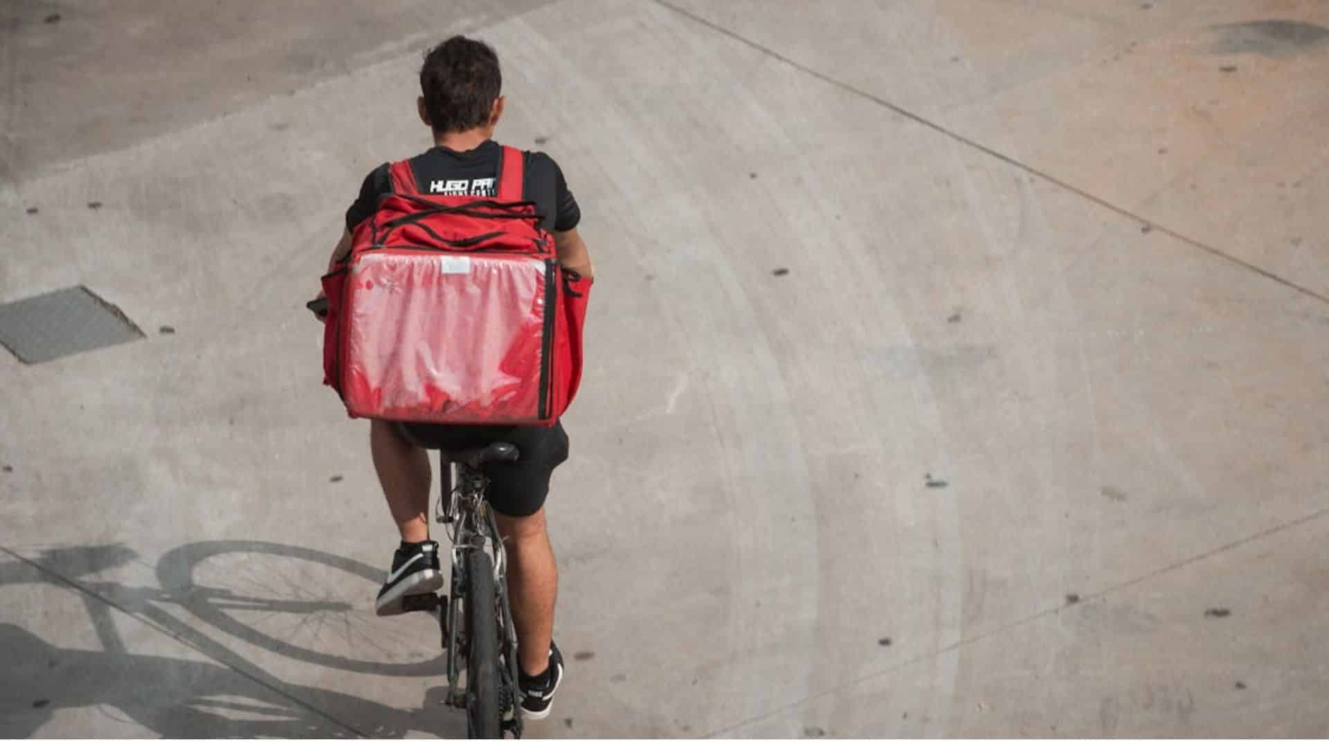 Delivery worker riding a bicycle with a red insulated bag on his back.