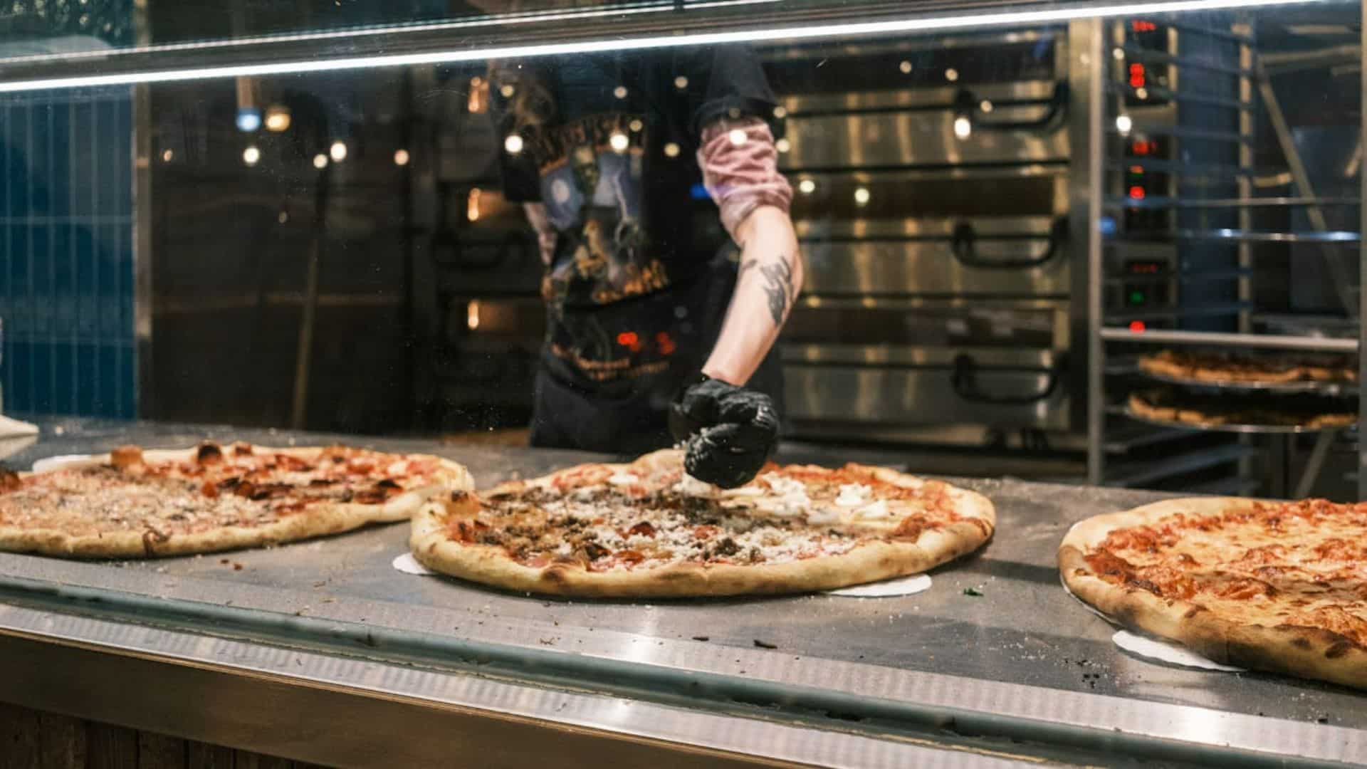 A person in a kitchen, wearing a black glove, prepares large pizzas behind a glass counter.