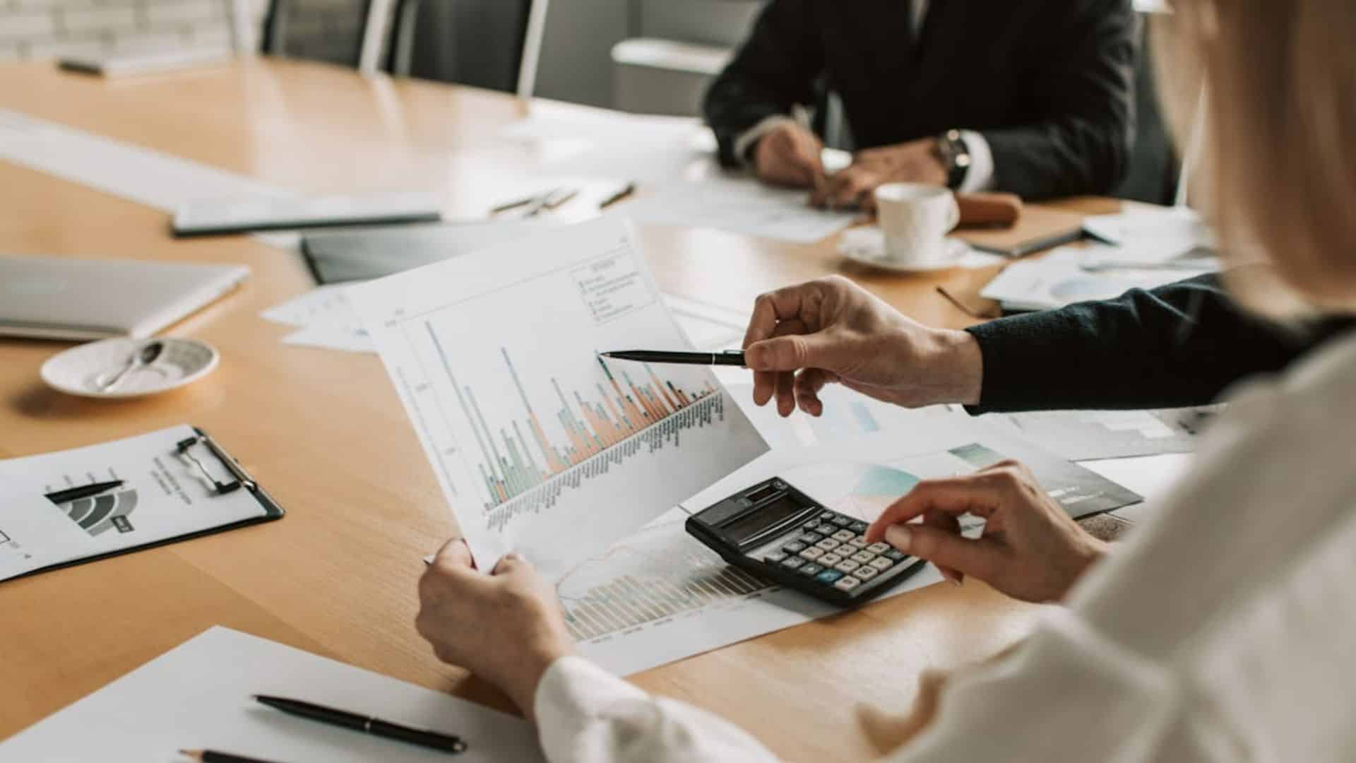 A group meeting with focused individuals, discussing a detailed bar chart. A calculator and papers are on the table.
