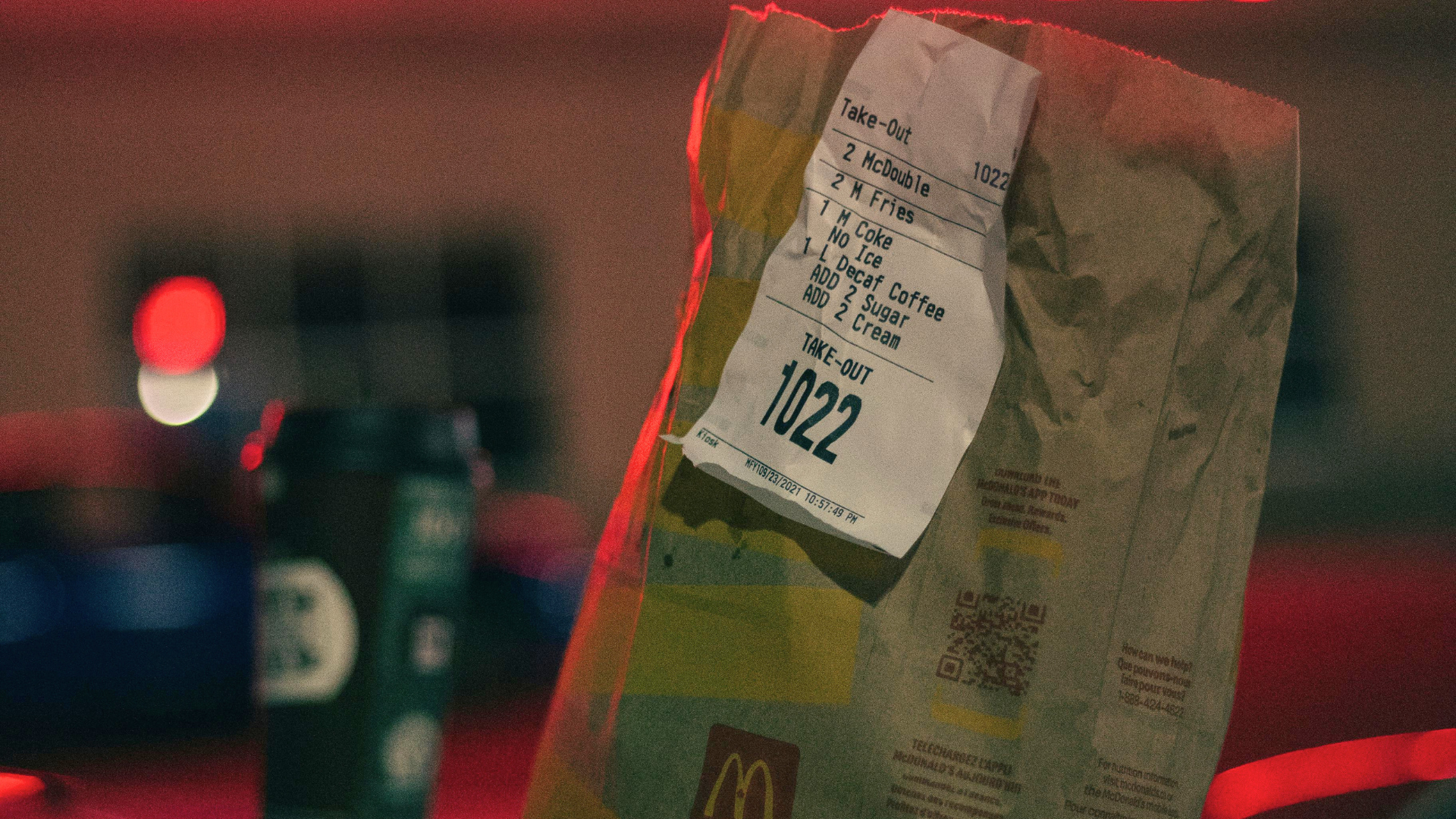 A McDonald's paper takeout bag with a detailed receipt attached sits on a dark surface at night under red neon lights.