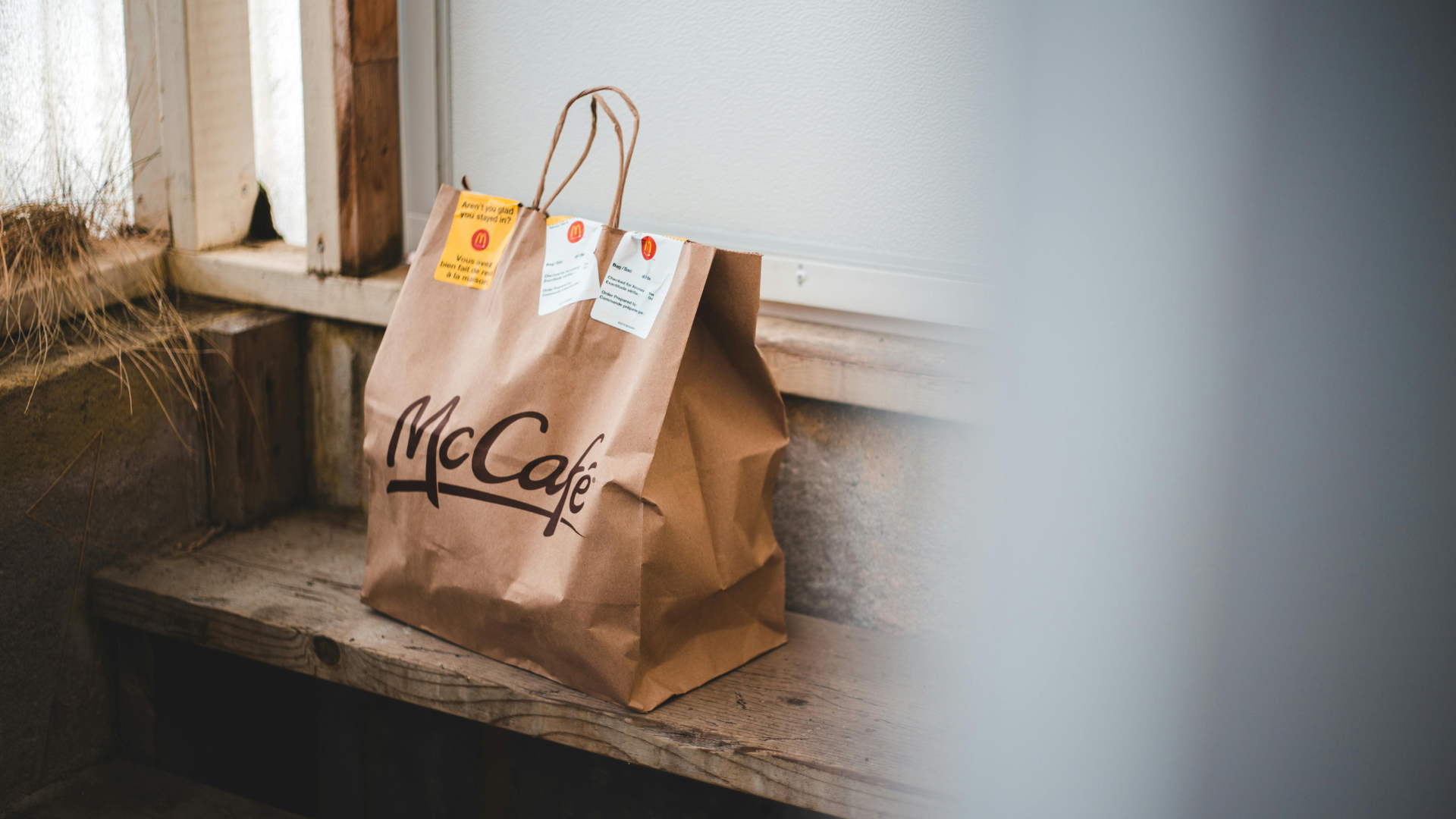 A brown McCafé paper delivery bag with white order receipts attached sits on a rustic wooden step.