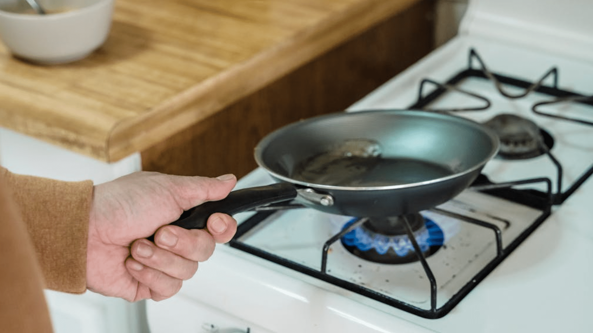 A close-up of a person's hand holding the handle of a small frying pan over a lit blue gas stove flame.