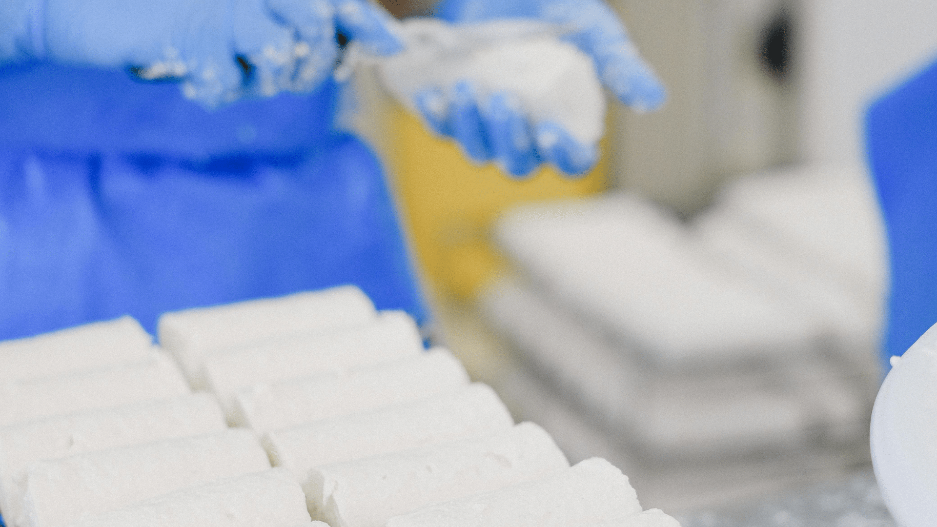 A person wearing blue gloves and a blue apron shapes soft, white cheese logs over a metal cooling rack in a commercial kitchen.
