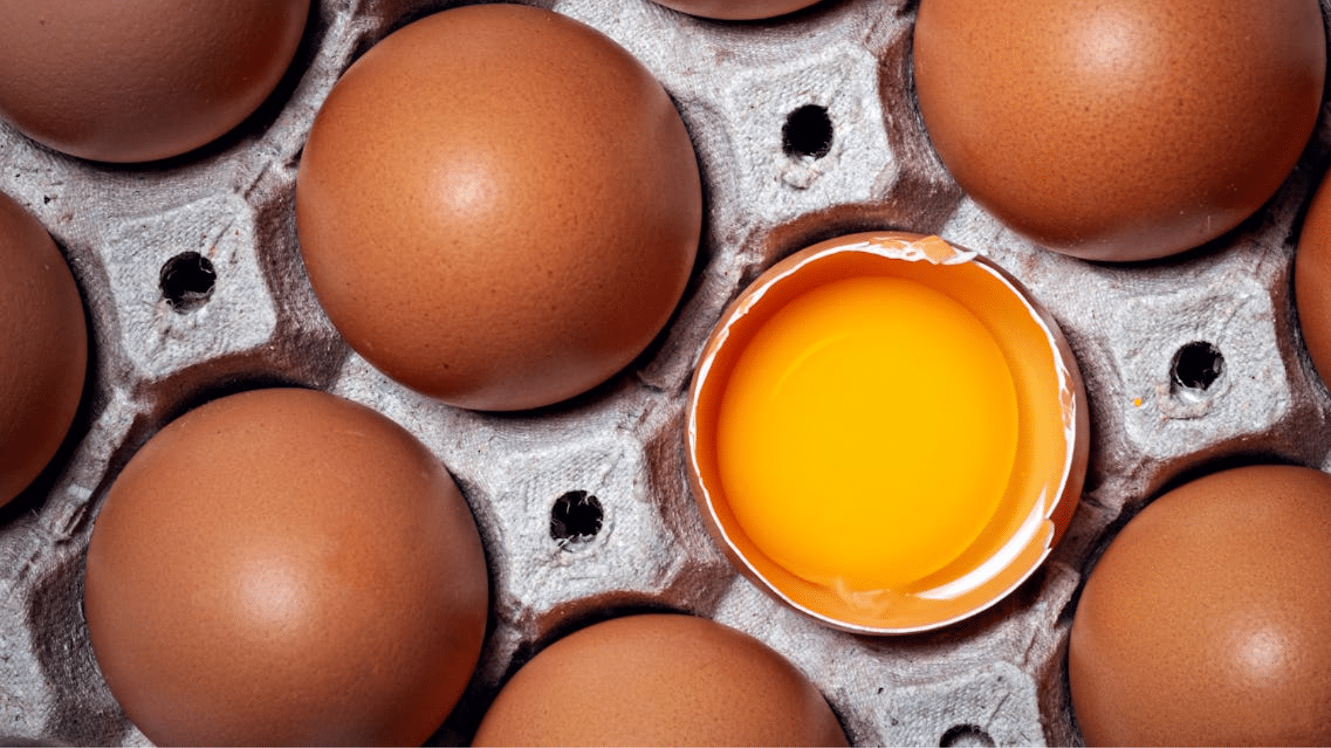 A top-down close-up of brown eggs in a carton, with one egg cracked open to reveal a bright, intact yellow yolk and clear albumen (egg white).