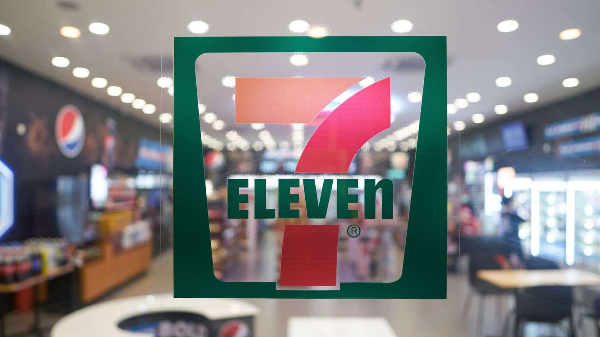 7-Eleven logo on a glass door of a brightly lit convenience store, displaying soft drinks and snacks.