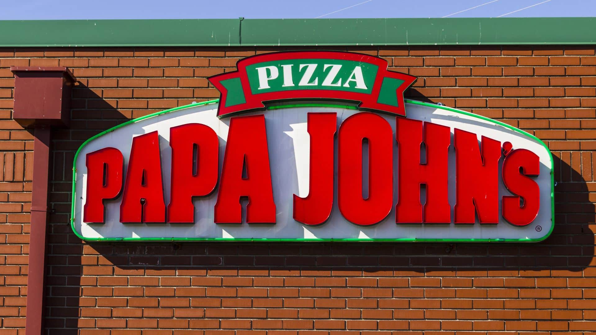 Sign for Papa John’s Pizza on a red brick wall, featuring bold red and green text and a green border.