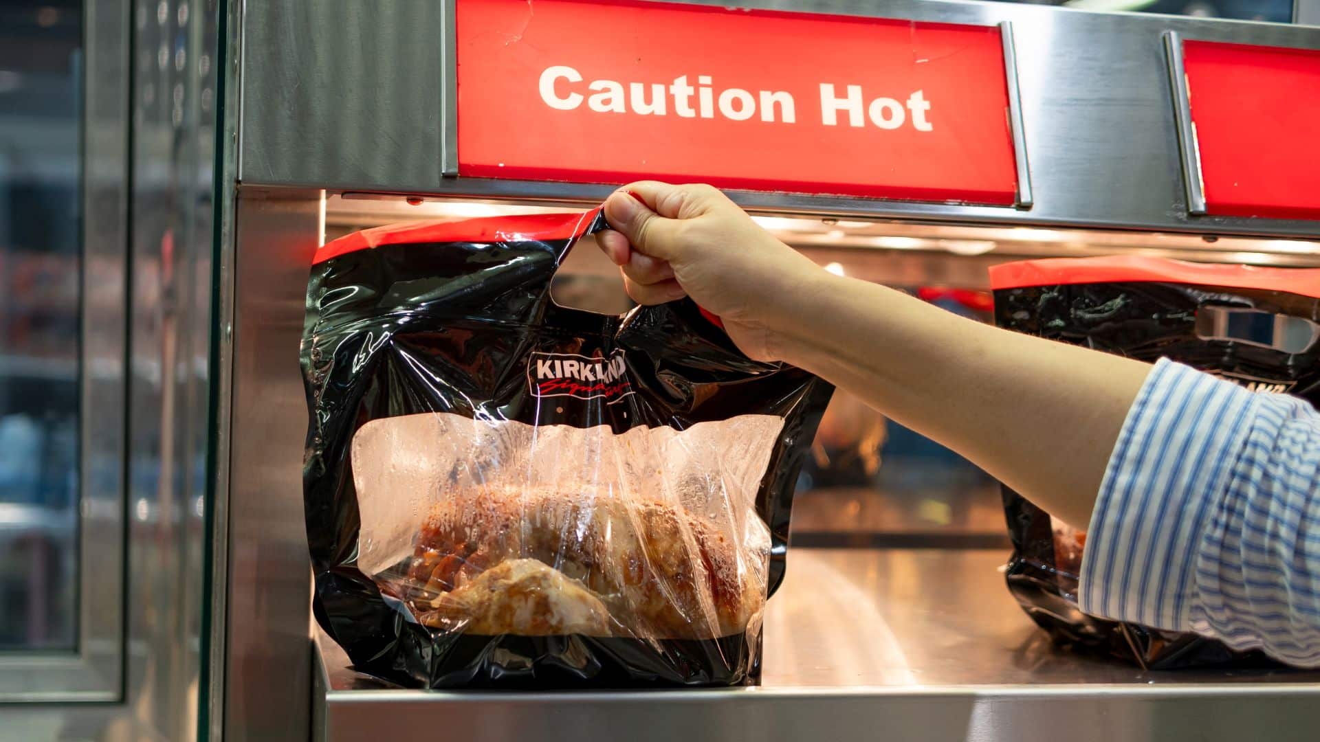 A hand picking up a Kirkland rotisserie chicken in a bag at a Costco food counter.