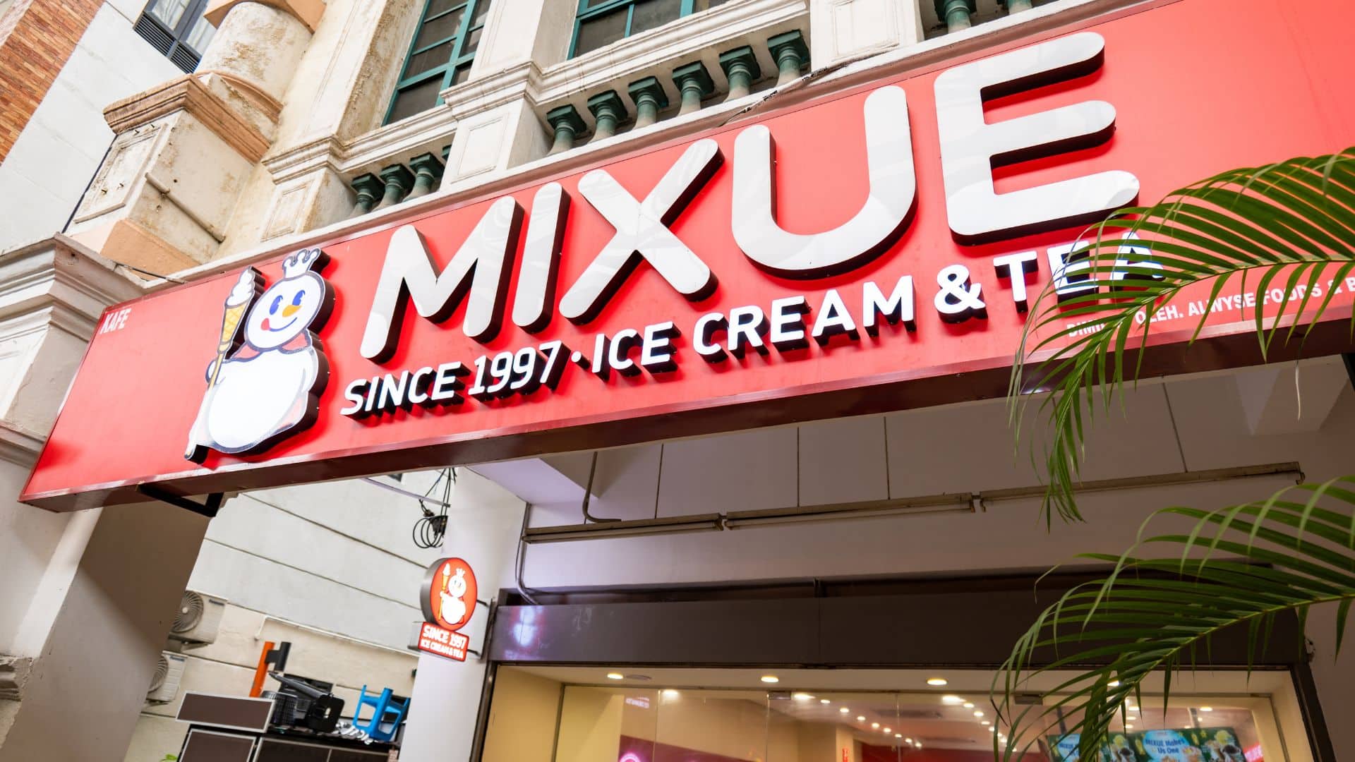 Red storefront sign of "Mixue" with a cartoon snowman logo, advertising ice cream and tea since 1997.