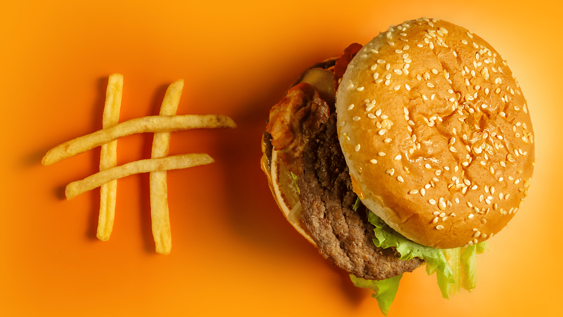 A top-down view of a gourmet burger with sesame bun, bacon, and lettuce, next to fries arranged in the shape of a hashtag.
