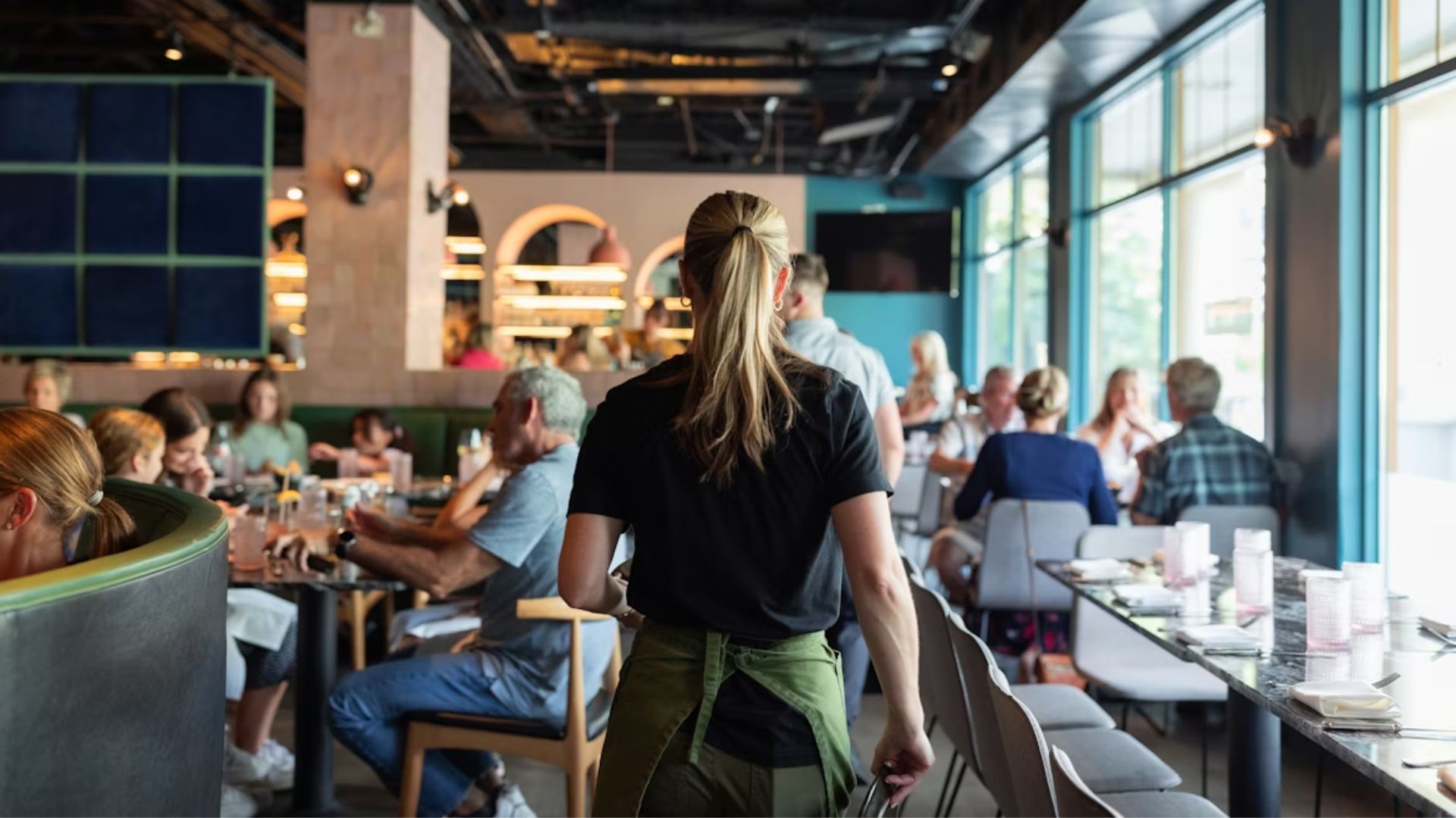 Server walking through busy restaurant dining room with seated guests at tables.