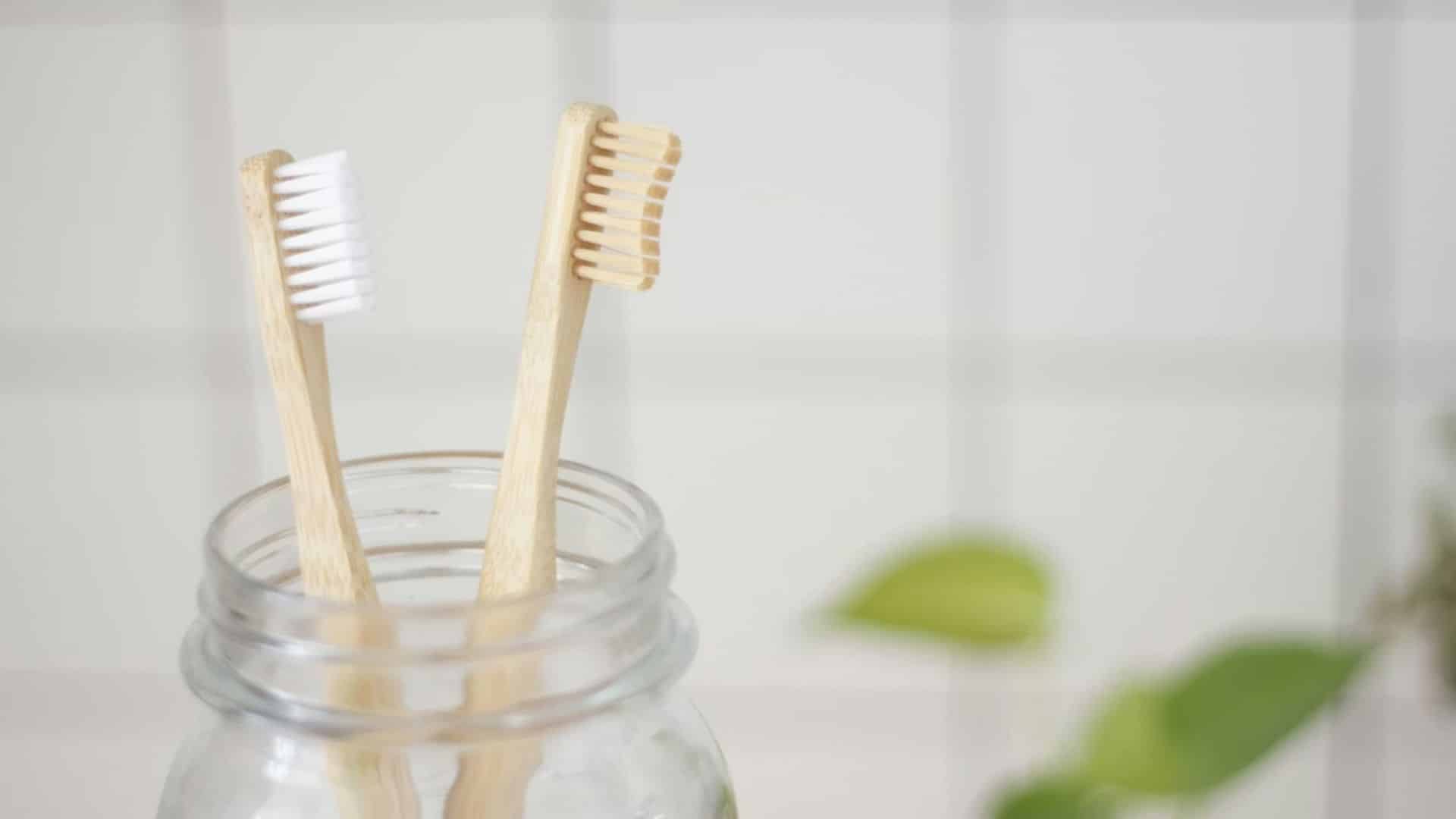 Two bamboo toothbrushes in a glass jar against a white tile background.