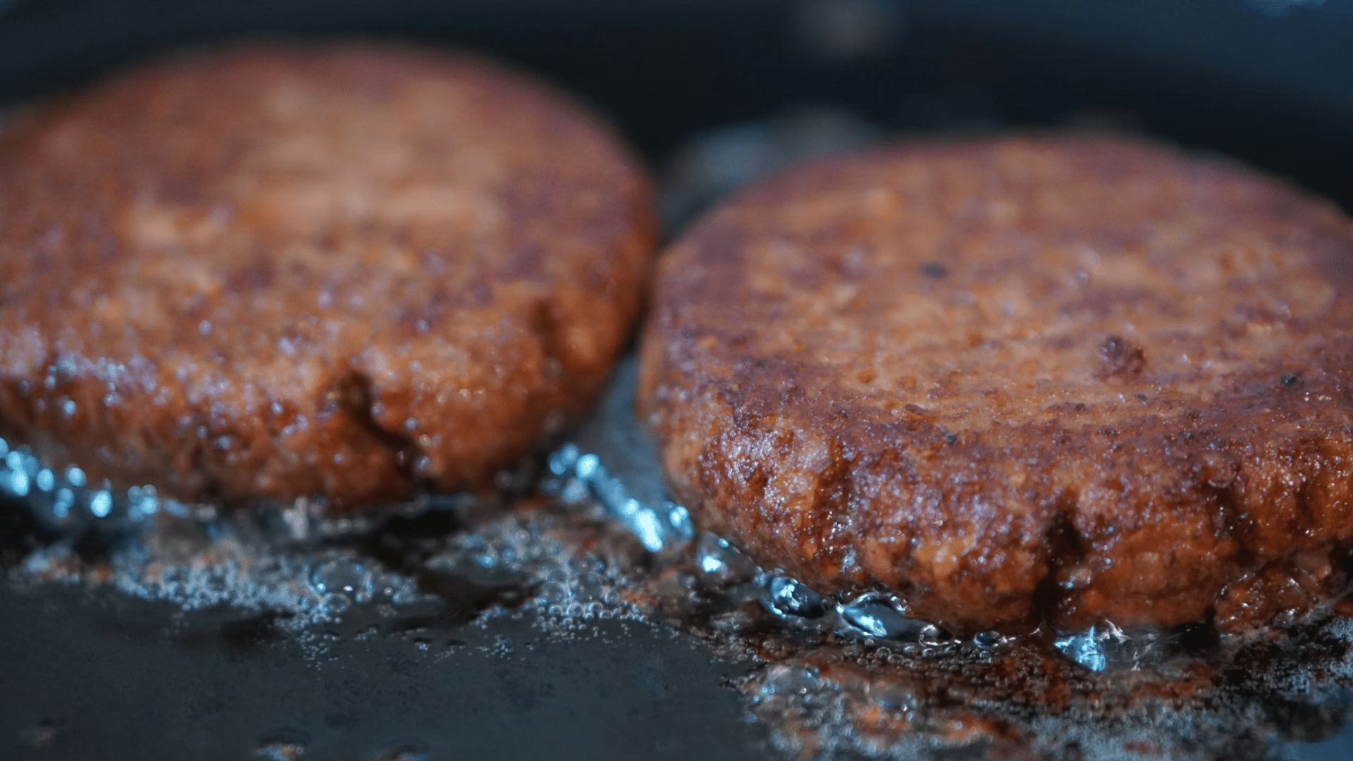 A close-up of two plant-based burger patties sizzling in a pan with grease bubbles.