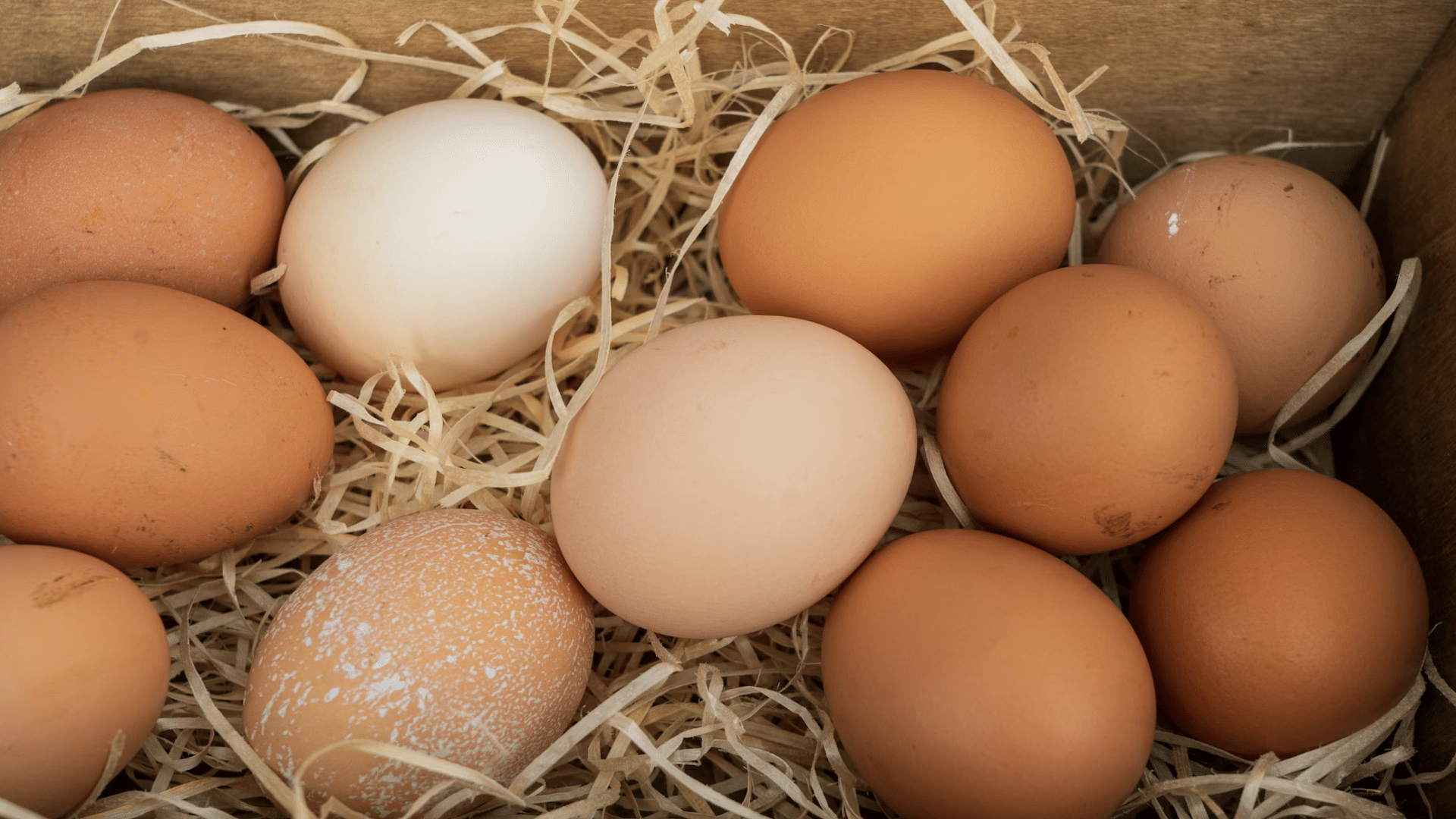 A top-down view of several brown and light-colored eggs resting on a bed of dry straw inside a wooden crate.