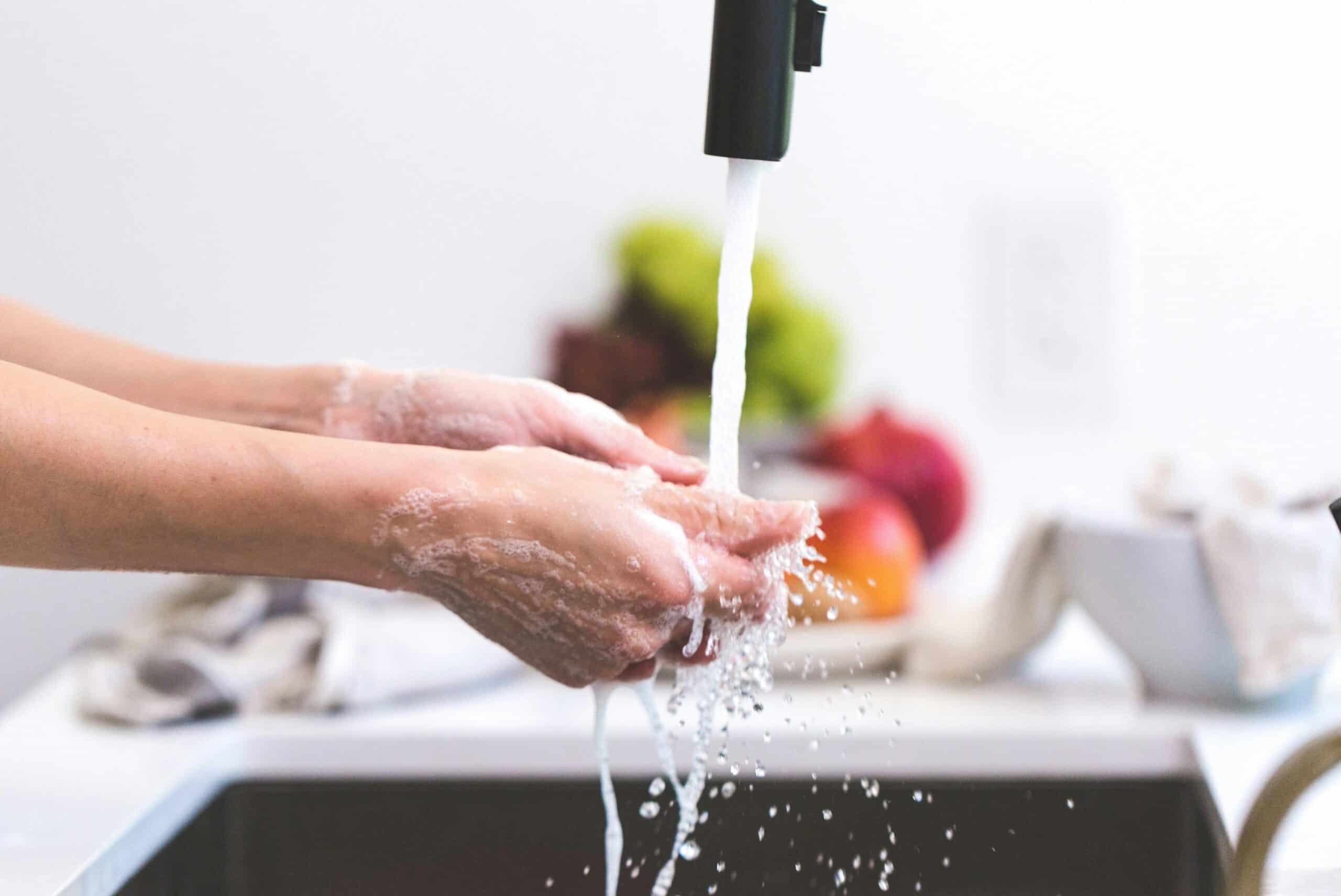 Person washing hands under running faucet with soap and water in a kitchen sink.