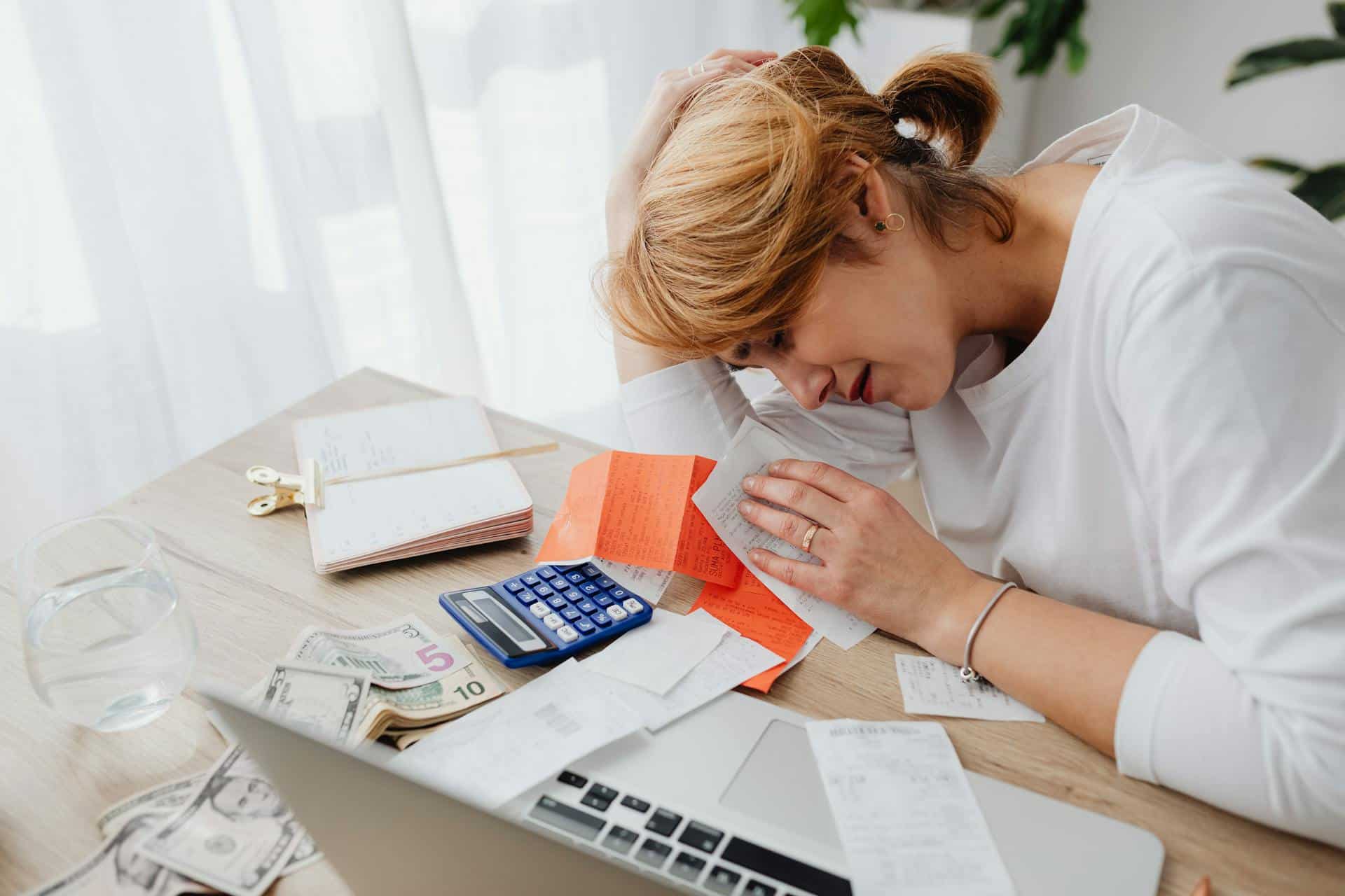 Woman reviewing bills and receipts with a calculator and laptop, holding her head in concern.
