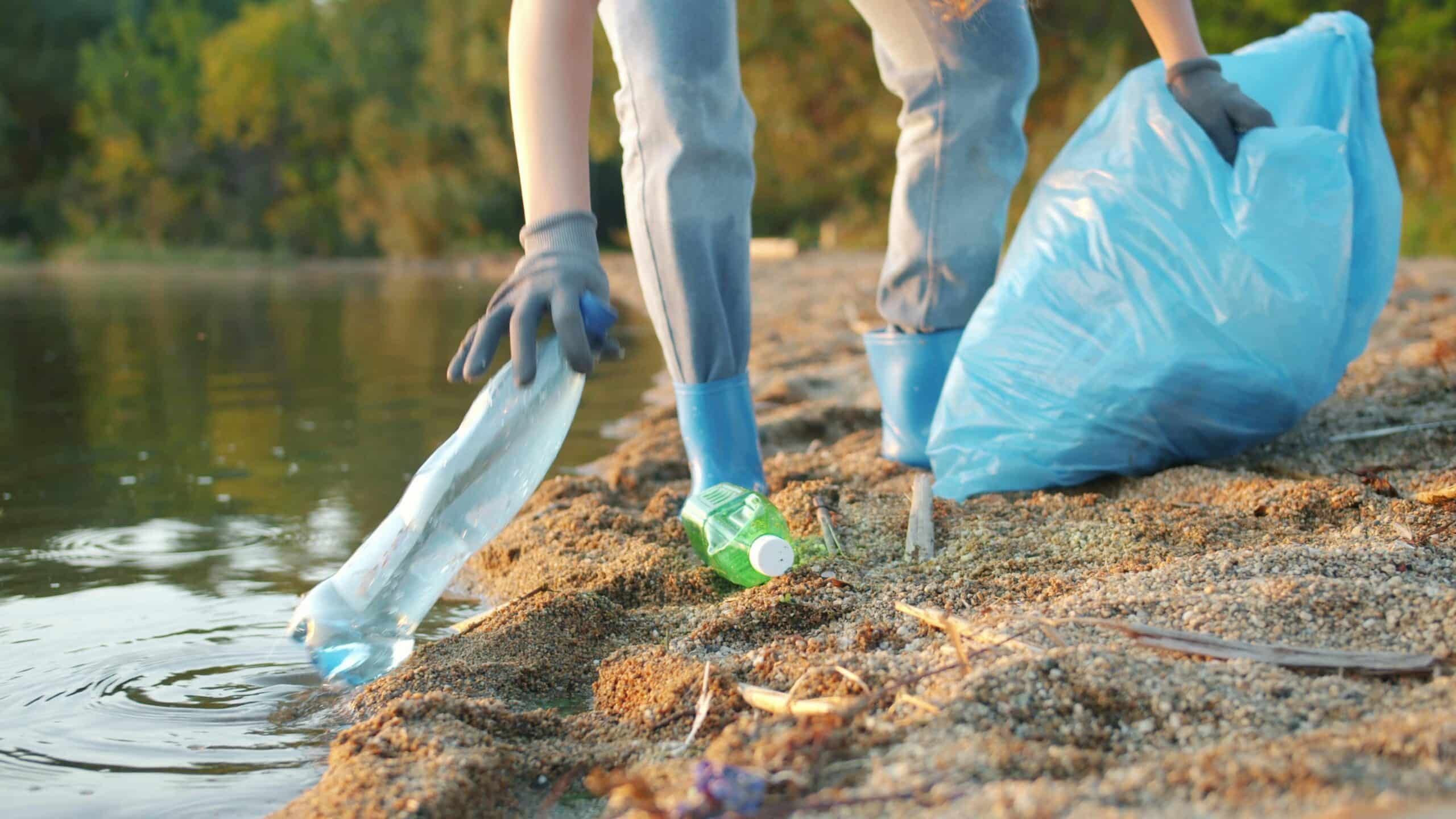Person wearing gloves collects plastic waste from a lakeshore, placing bottles into a trash bag.
