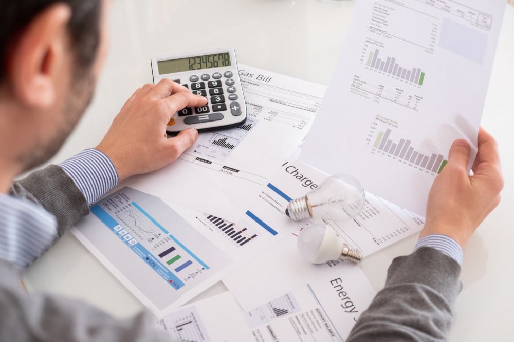 Man using a calculator while reviewing energy bills and charts with light bulbs on the table.