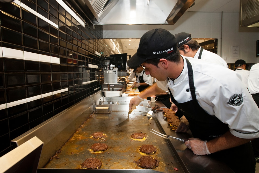 Steak ’n Shake employees cooking steakburger patties on a flat-top grill in a restaurant kitchen.