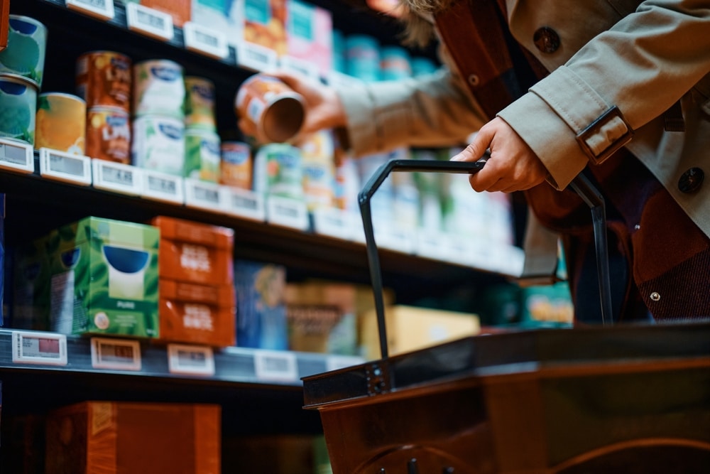 Shopper holding a can with a basket in hand in front of shelves stocked with packaged goods.