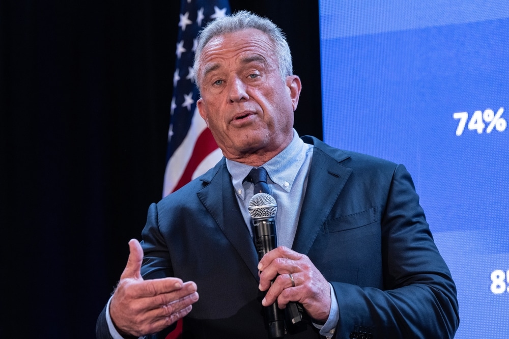 Robert F. Kennedy Jr. speaking into a microphone at a public event with a U.S. flag in the background.