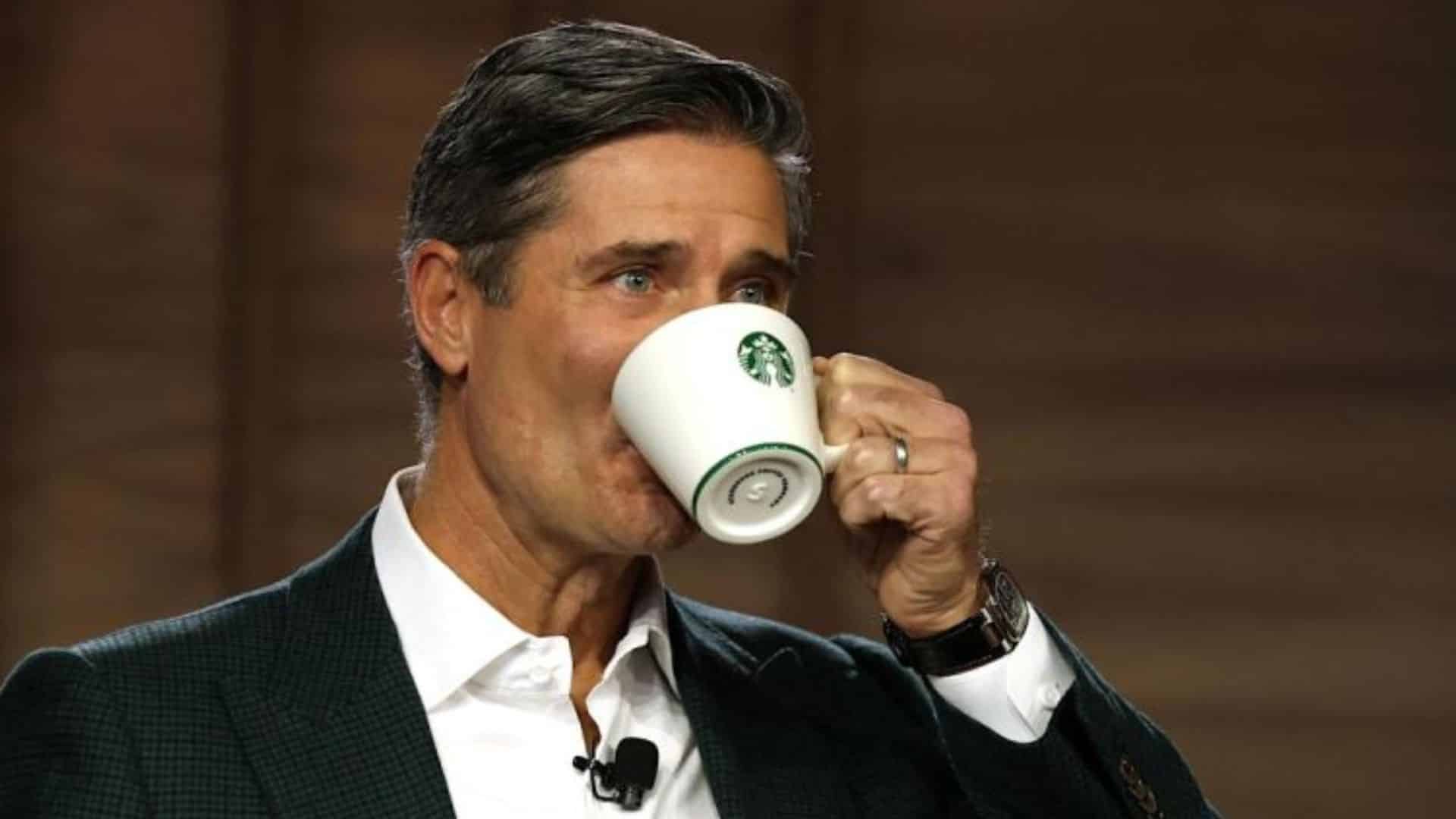 Starbucks CEO Brian Niccol speaking on stage while drinking from a white Starbucks mug during a public appearance.