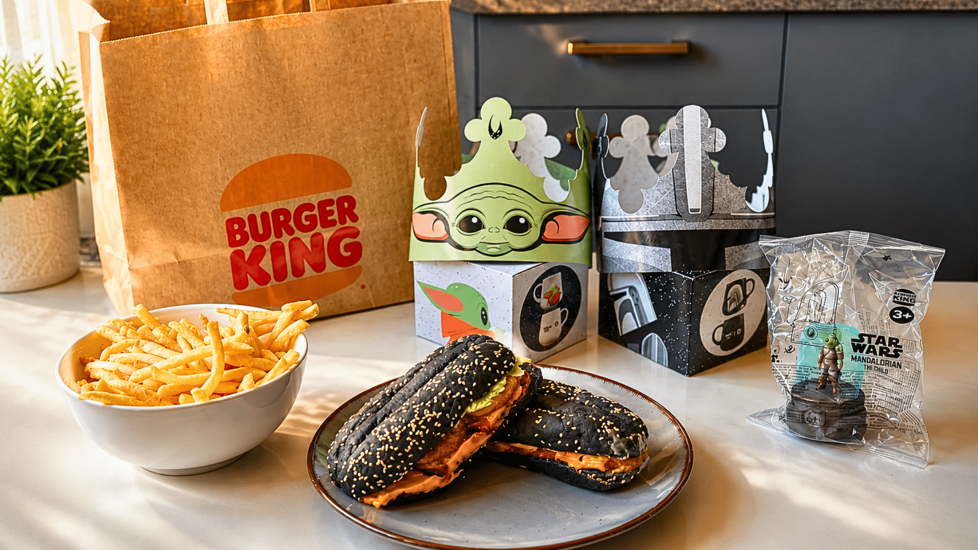 A well-lit kitchen counter display featuring a Burger King paper bag and a variety of The Mandalorian-themed items; includes two cardboard crowns—one green with Grogu's face and one silver resembling a Mandalorian helmet—two black-bun burgers on a grey plate, a bowl of fries, and a packaged Star Wars toy.